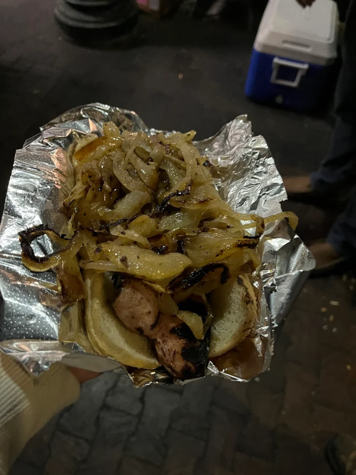 A grilled sausage in a bun topped with a generous amount of sautéed onions, served on aluminum foil and held in someone's hand. The background is dimly lit, suggesting a nighttime setting.