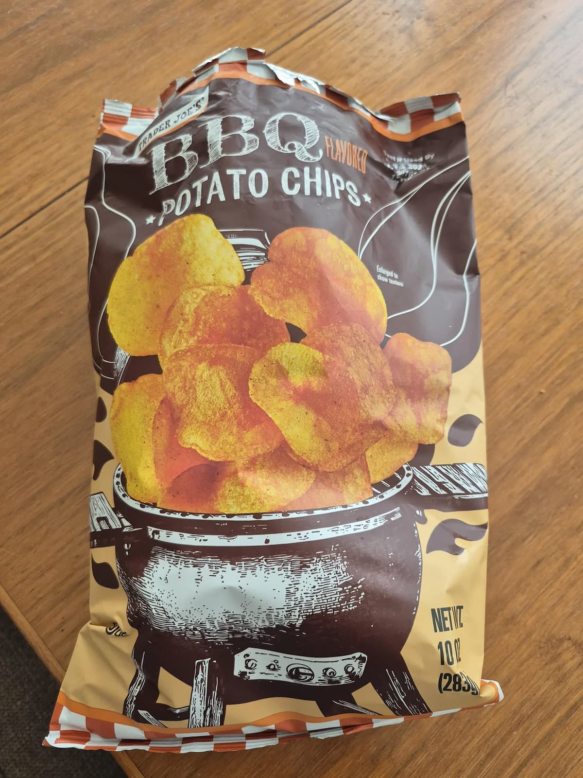 A bag of Trader Joe's BBQ flavored potato chips sits on a wooden table. The packaging shows an illustration of potato chips above a charcoal grill, with brown and orange colors dominating the bag design. Net weight: 10 oz.