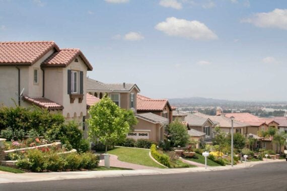 A middle class suburban neighborhood with large, beige houses featuring red-tiled roofs, well-kept gardens, green lawns, and a sloping street under a partly cloudy sky. City buildings are visible in the distant background.