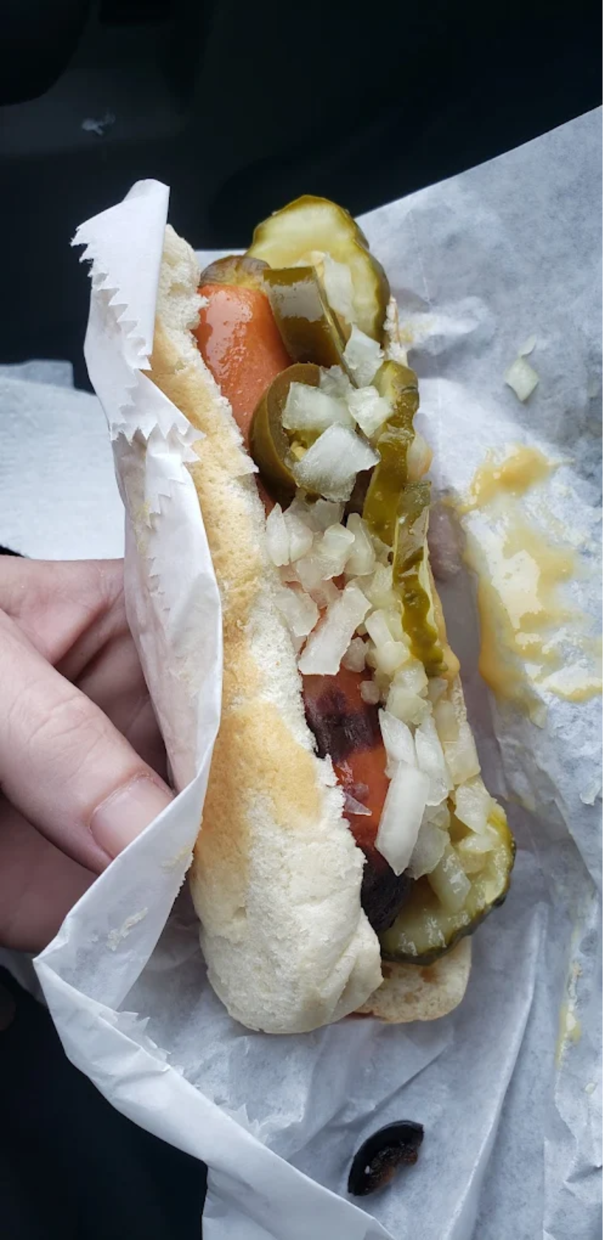 A hand holds a hotdog in a bun topped with chopped onions, sliced pickles, and jalapeños, wrapped partly in white paper with some mustard visible on the wrapping.