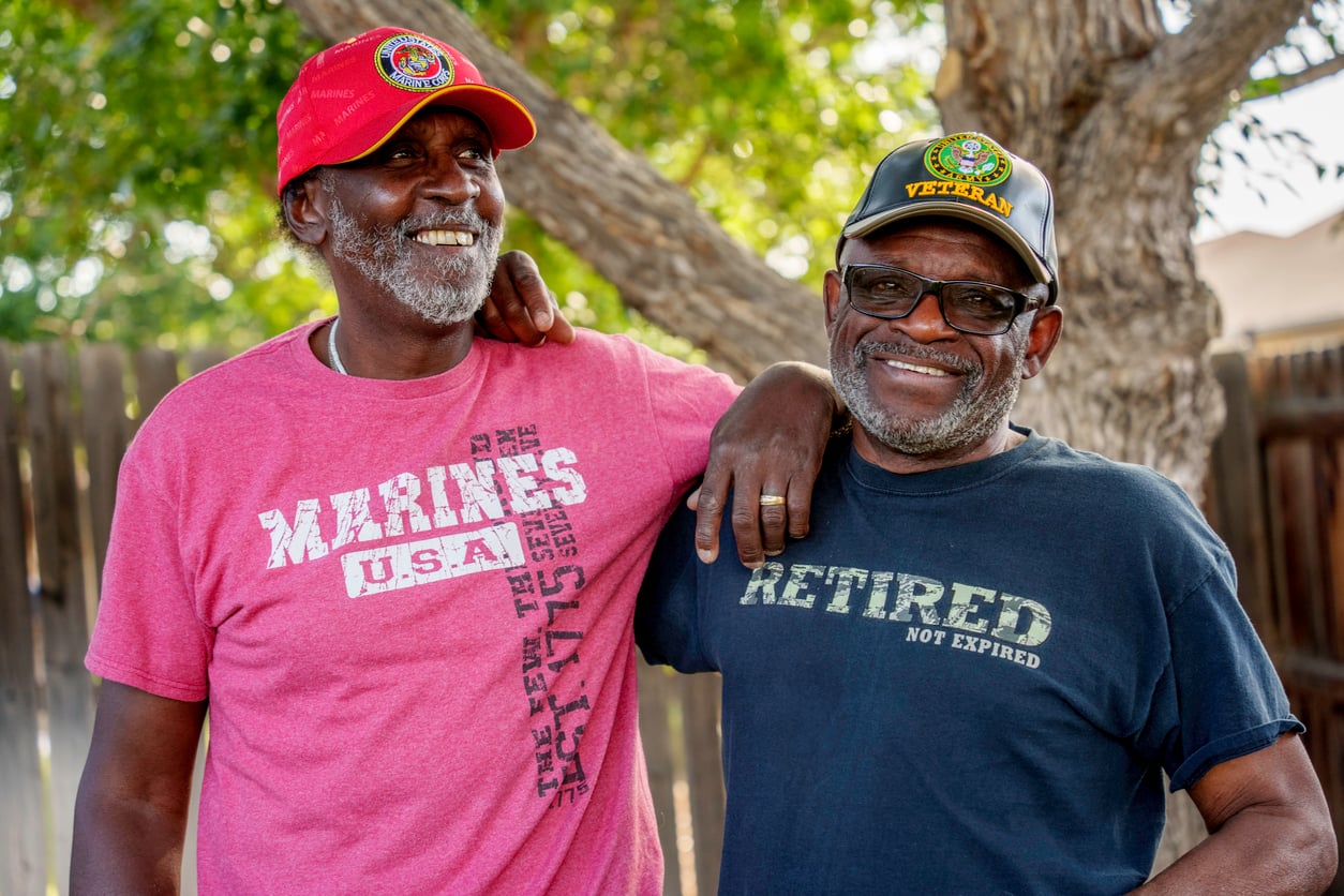 Two older men stand outdoors smiling. One wears a red "MARINES U.S.A." shirt and cap, and the other a black "RETIRED" shirt and "VETERAN" cap—enjoying good company and the camaraderie that comes from sharing veteran benefits.