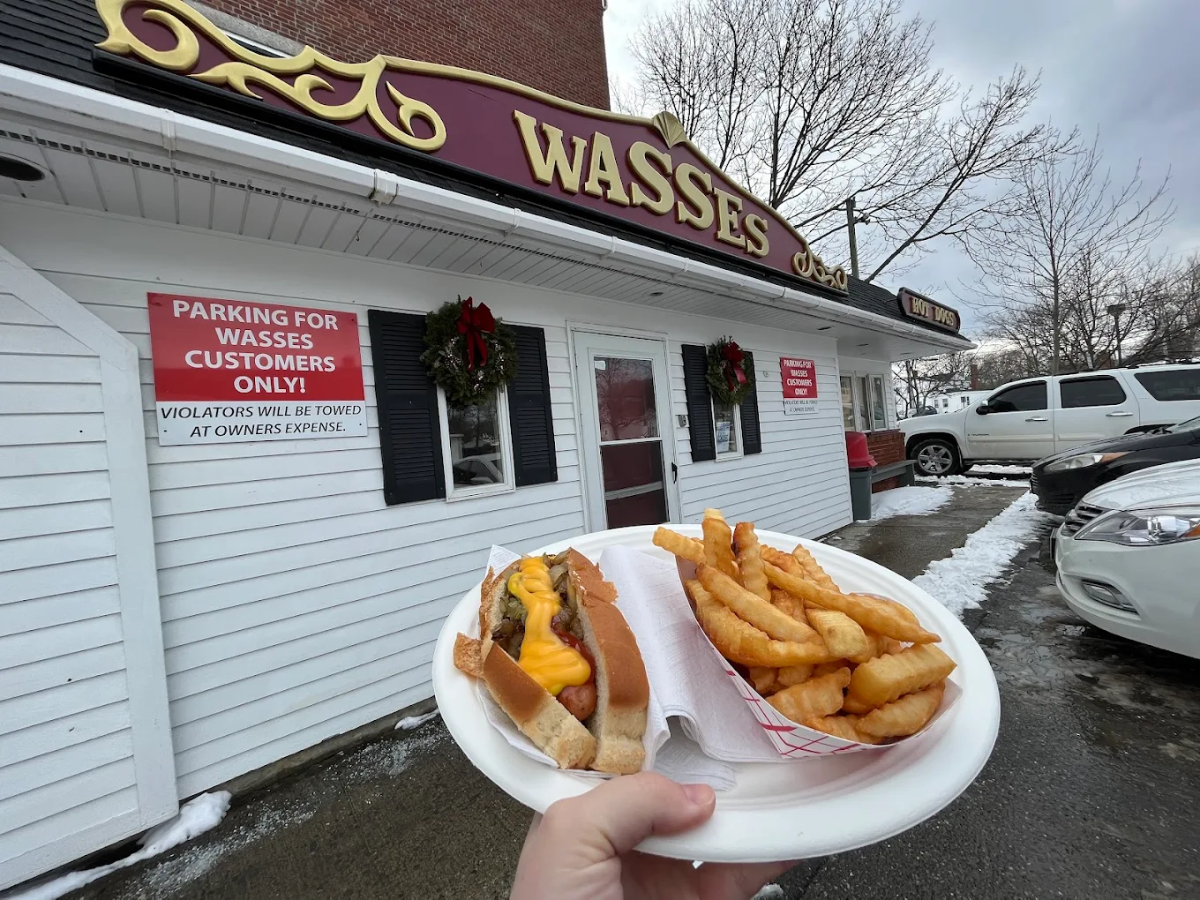 A hand holds a tray with a hot dog topped with cheese and crinkle-cut fries in front of Wasses restaurant on a cloudy winter day with parked cars and snow on the ground.