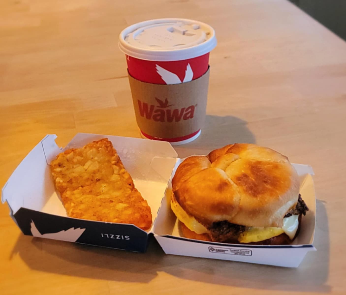 A Wawa-branded cup of coffee sits on a table next to a breakfast sandwich with egg, cheese, and meat on a bun, and a golden hash brown in a cardboard tray.