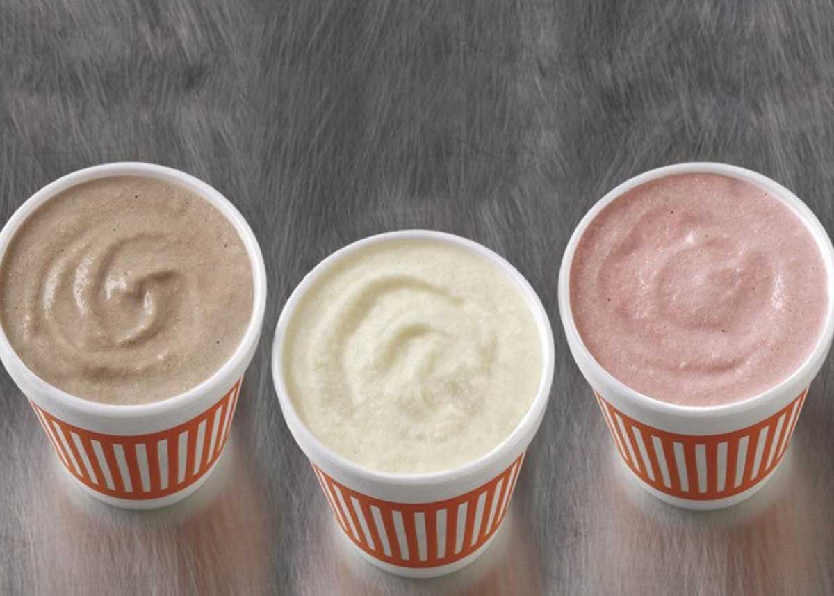Three cups of milkshakes—chocolate, vanilla, and strawberry—are arranged side by side on a gray surface. Each cup has an orange and white striped design.