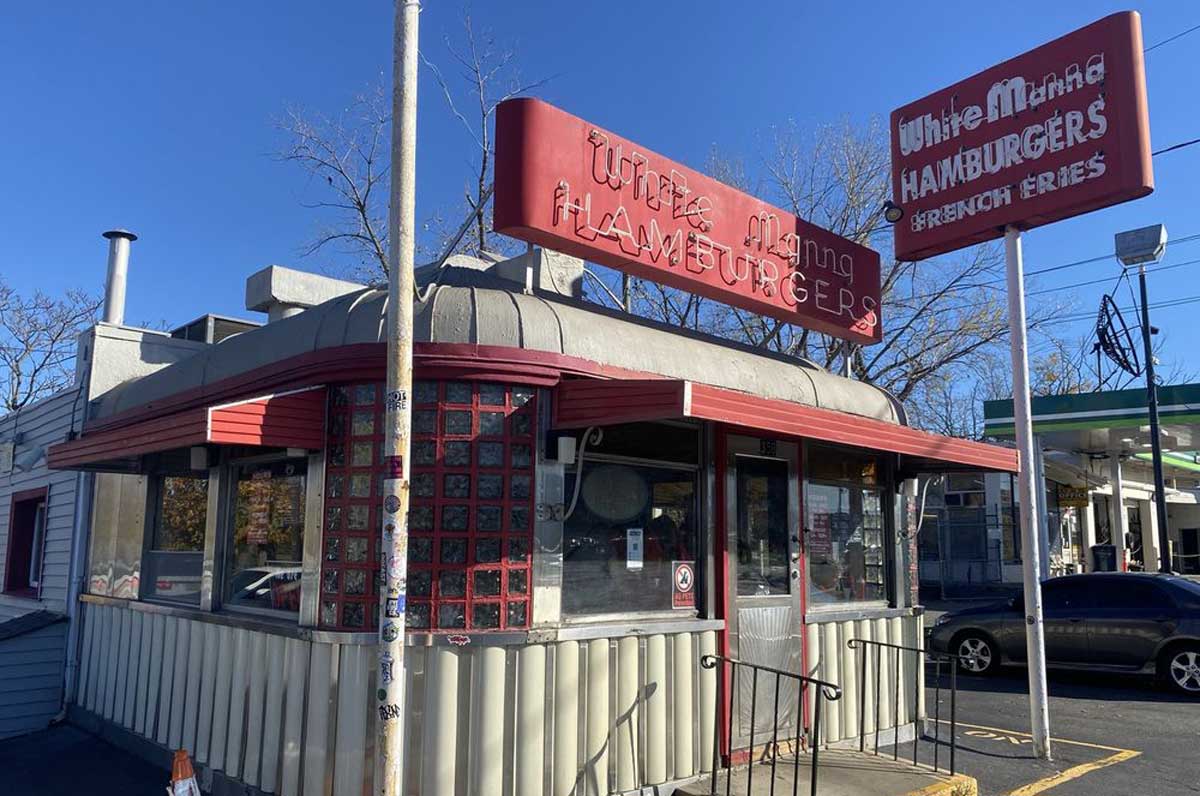 A vintage-style diner with red trim and signs reading "White Manna HAMBURGERS" and "French Fries" stands on a sunny day, featuring large windows and a metal exterior, capturing the timeless charm of old-school diners in America.