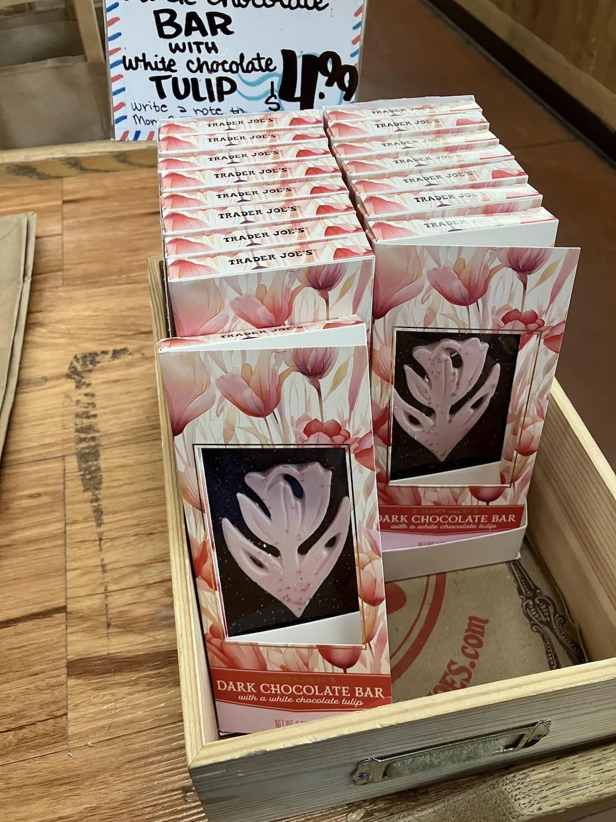 A wooden crate displays several Trader Joe’s dark chocolate bars, each with a visible pink and white tulip-shaped chocolate inside floral packaging. A handwritten sign above reads, "Dark Chocolate Bar with White Chocolate Tulip $1.99".