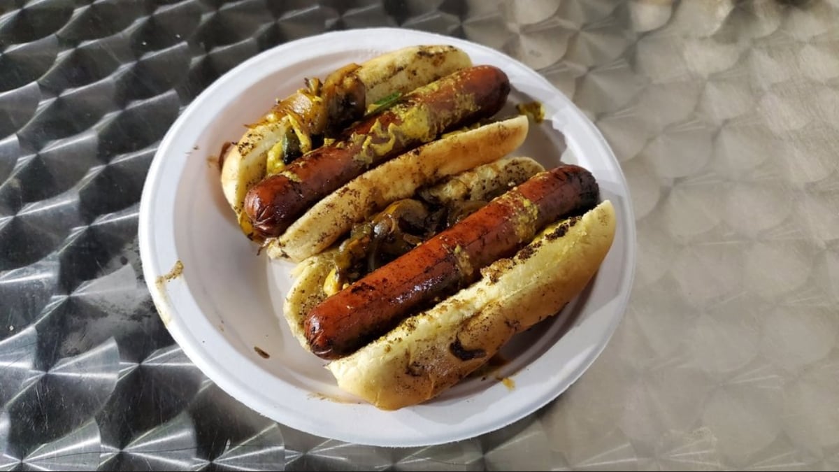Three grilled hot dogs in buns, topped with sautéed onions and mustard, are served on a white disposable plate placed on a metallic textured surface.