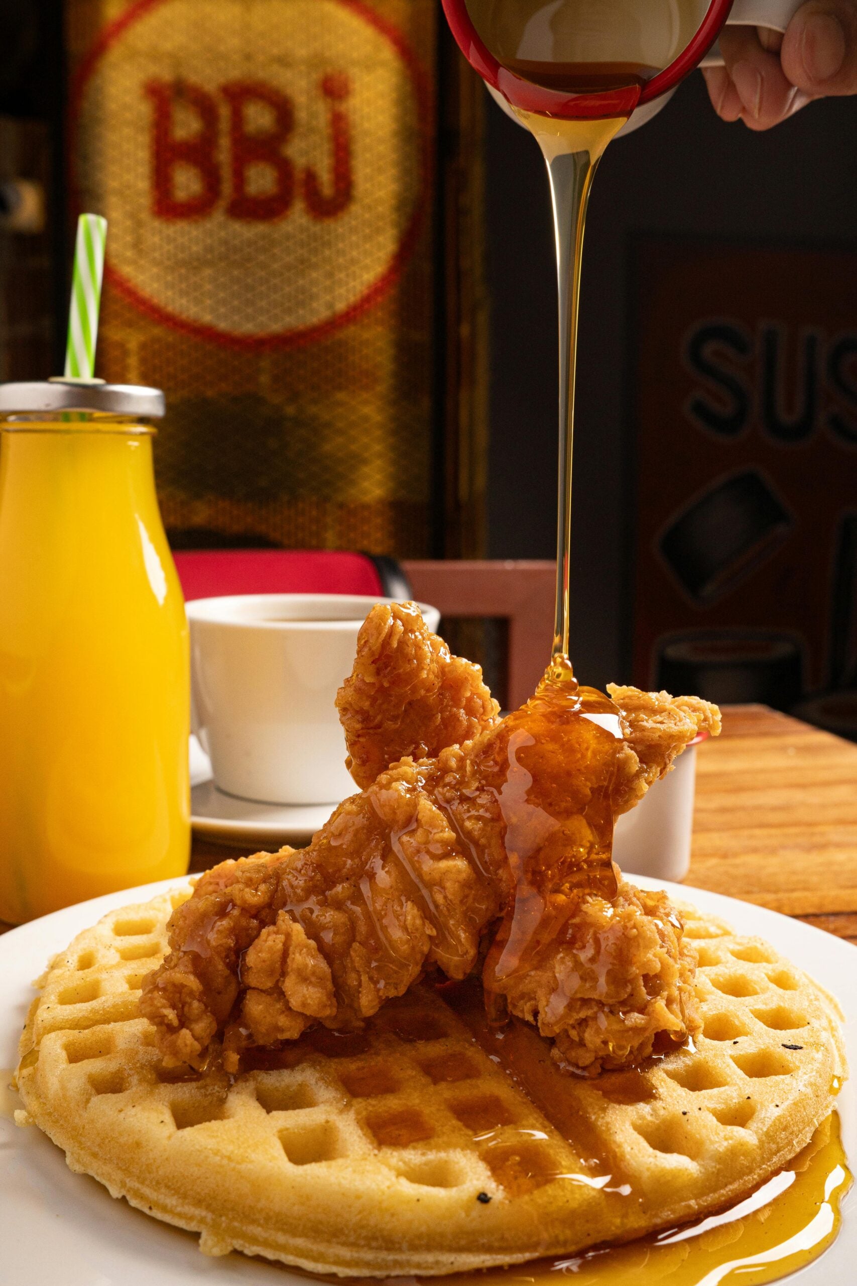 A plate of chicken and waffles is topped with fried chicken strips, while syrup is being poured over them. A glass bottle of orange juice and a white coffee cup sit in the background.