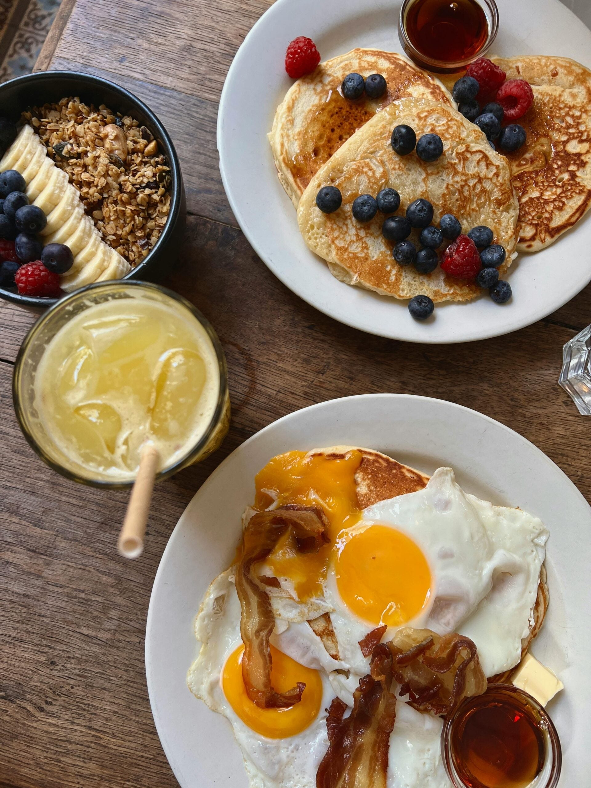 Breakfast spread on a wooden table with pancakes topped with berries and syrup, a bowl of granola with banana slices, a plate of fried eggs and bacon, and a glass of yellow juice with a straw.