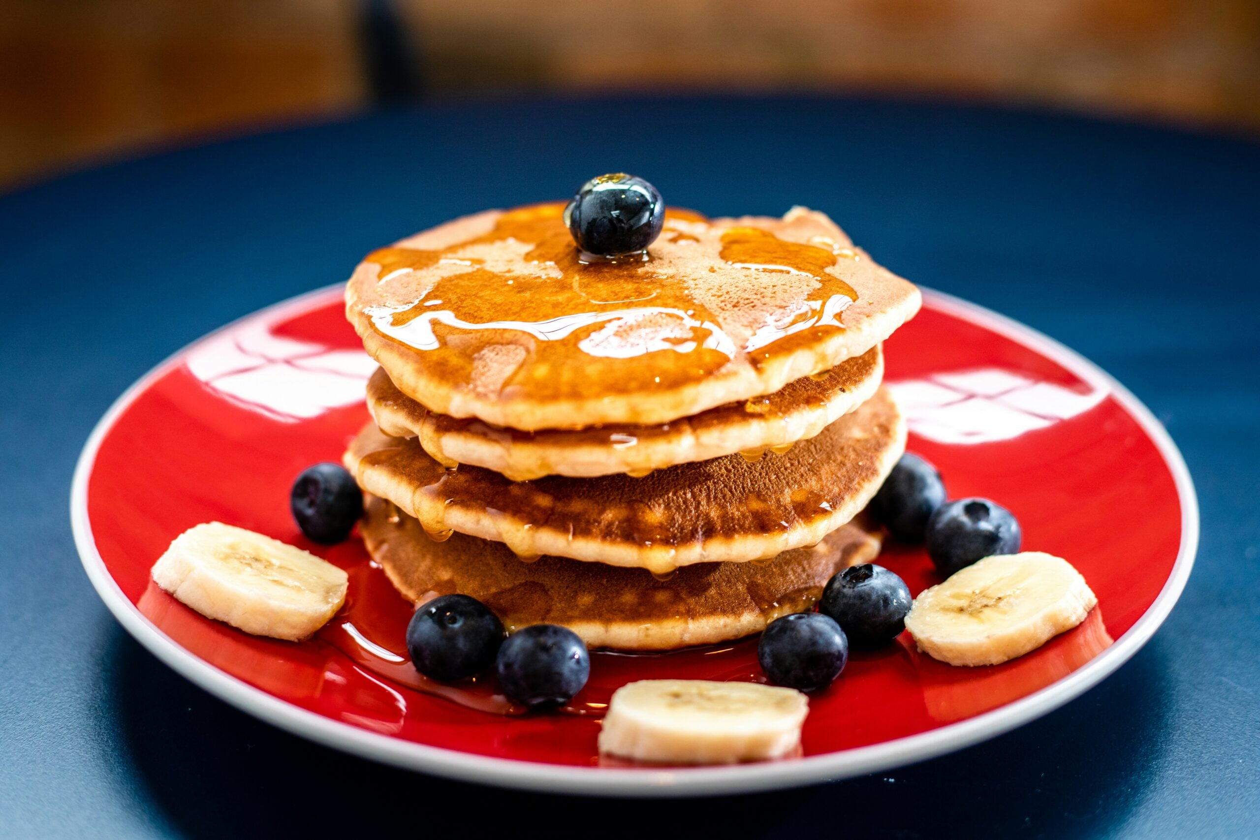 A stack of pancakes topped with a blueberry and drizzled with syrup, served on a red plate with blueberries and banana slices on a dark surface.