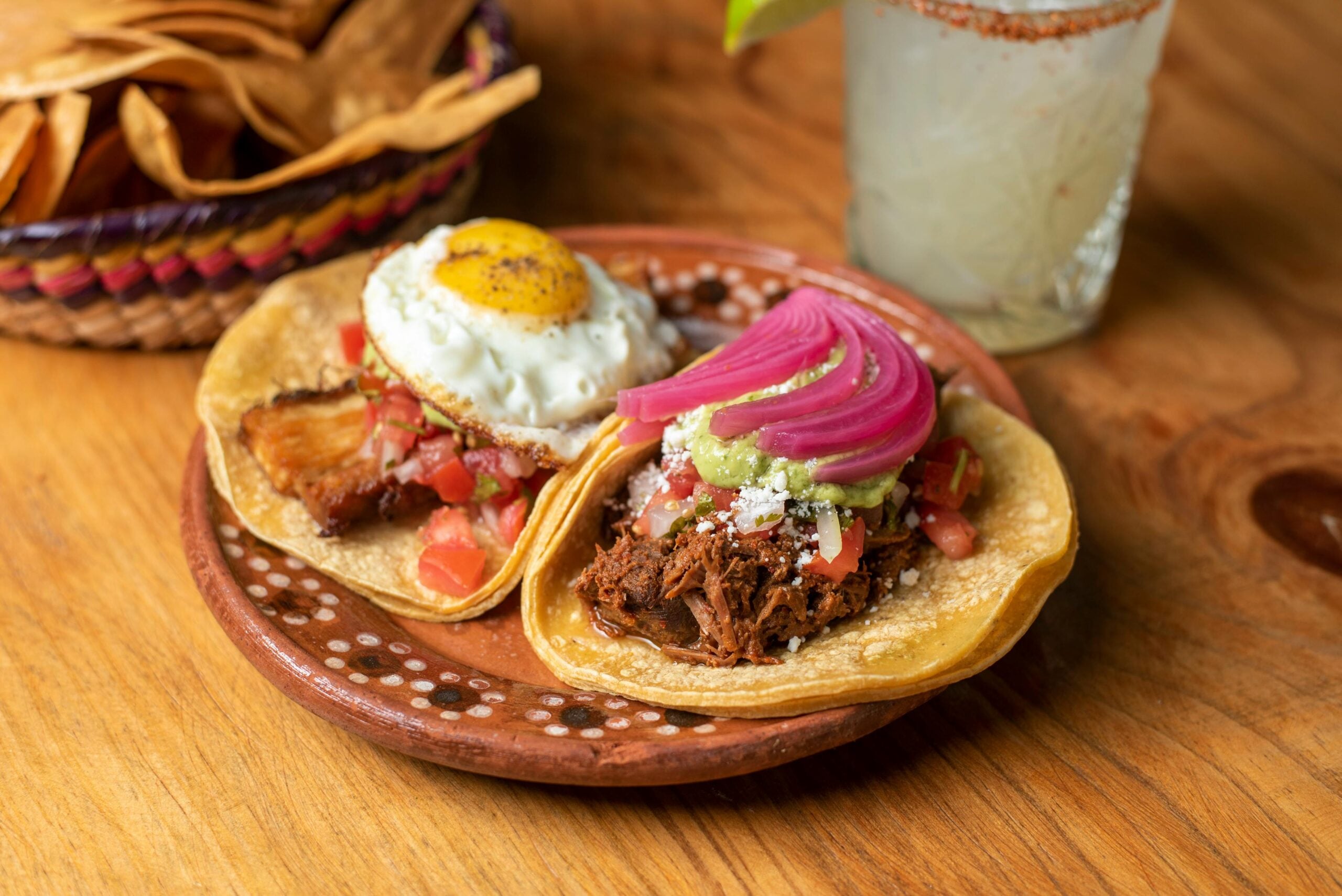 Two tacos on a decorative plate: one topped with shredded meat, diced tomatoes, pickled red onions, guacamole, and cheese; the other with roasted pork, pico de gallo, and a fried egg. Drink and tortilla chips in the background.