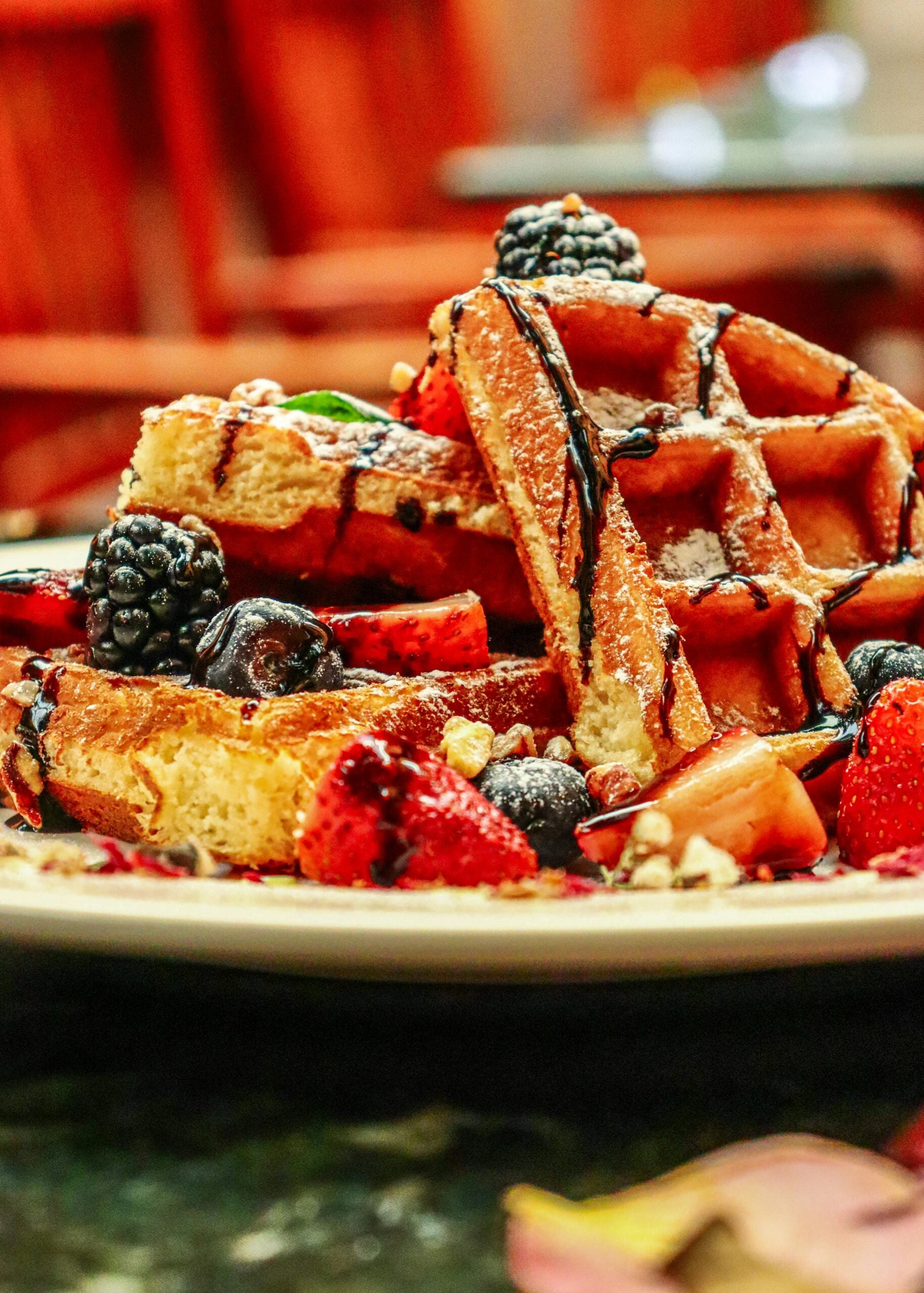A plate of golden waffles topped with fresh strawberries, blueberries, and blackberries, drizzled with chocolate sauce and sprinkled with powdered sugar and nuts.