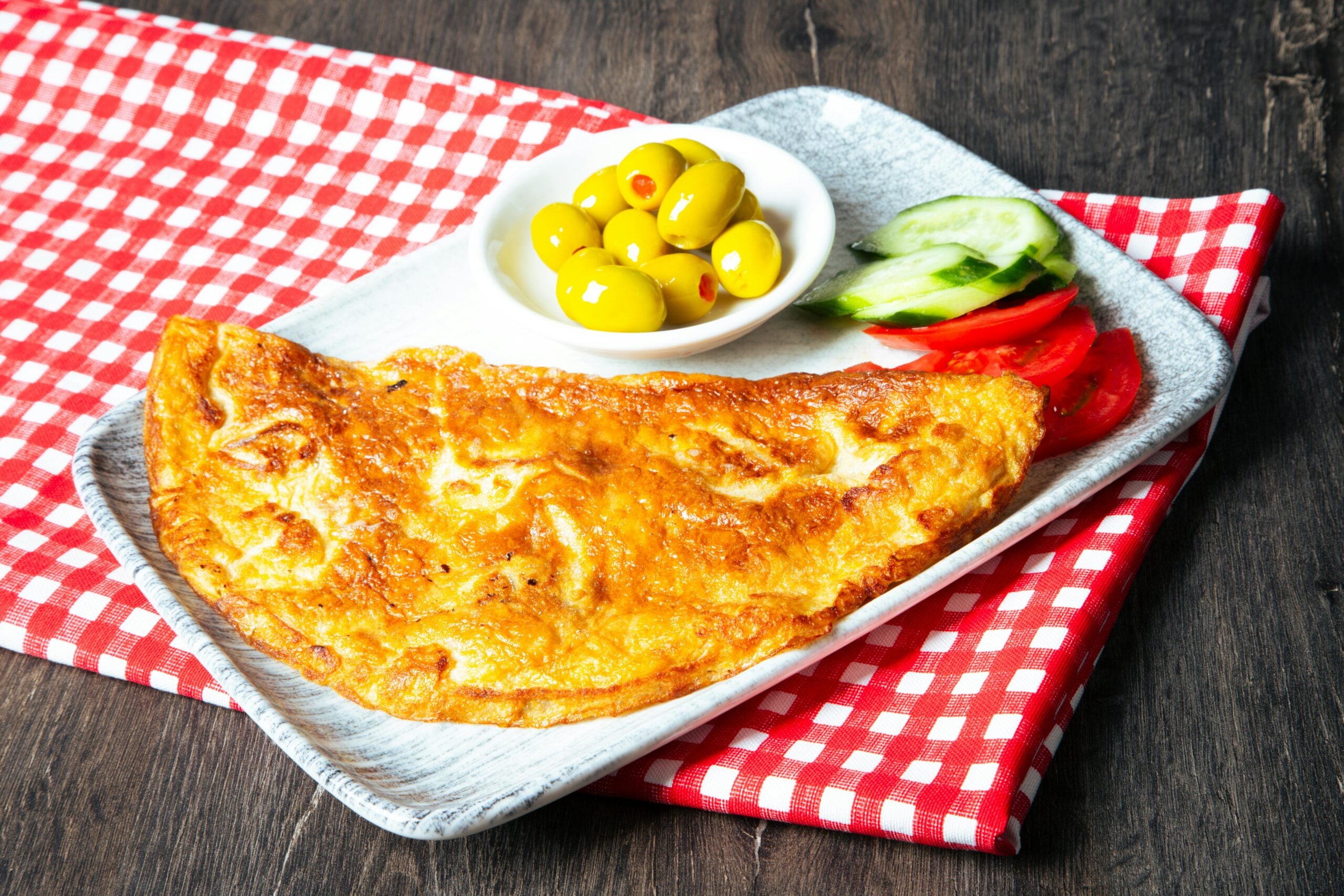 A golden omelette on a rectangular plate with a side of green olives in a small bowl, sliced cucumber, and tomato, all placed on a red and white checkered cloth on a dark wooden table.