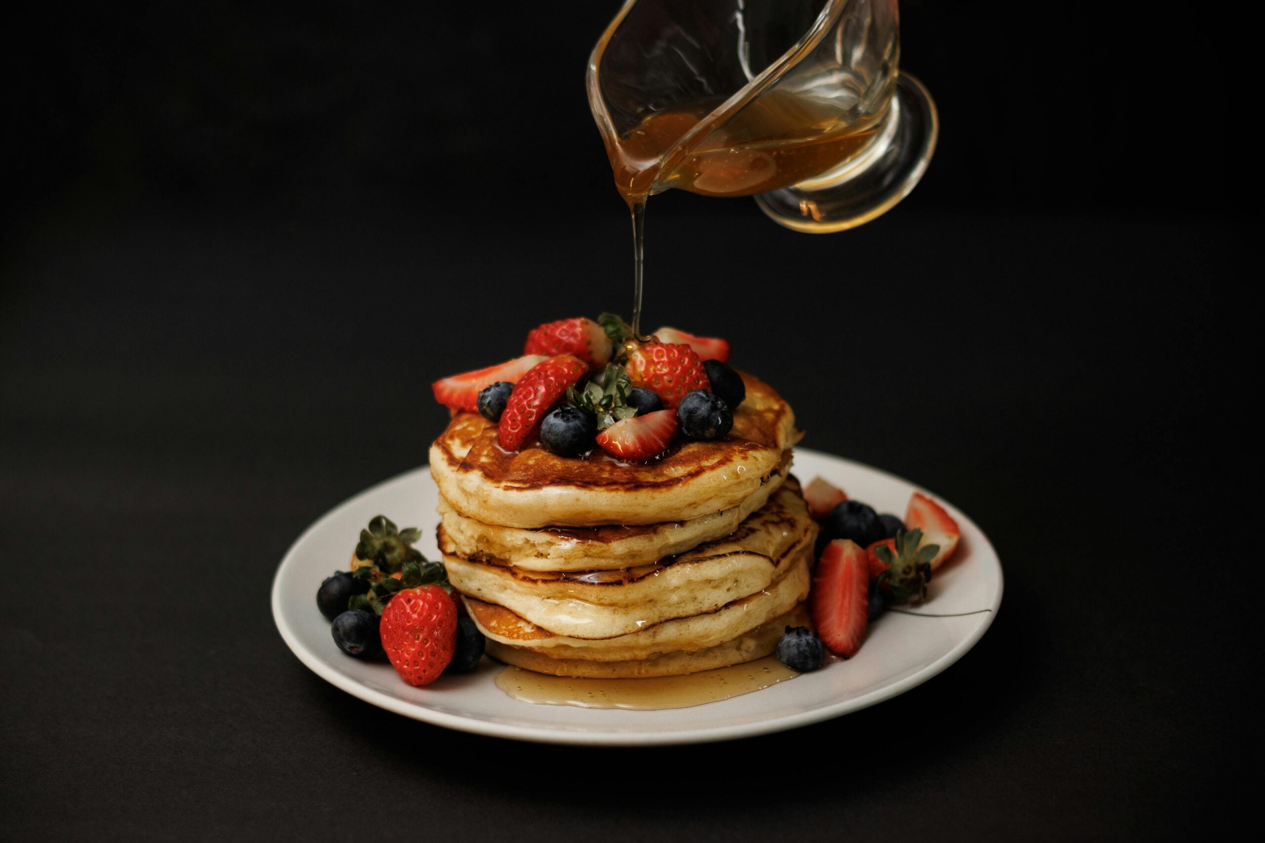 A stack of fluffy pancakes topped with strawberries and blueberries on a white plate, with syrup being poured over them, set against a dark background.