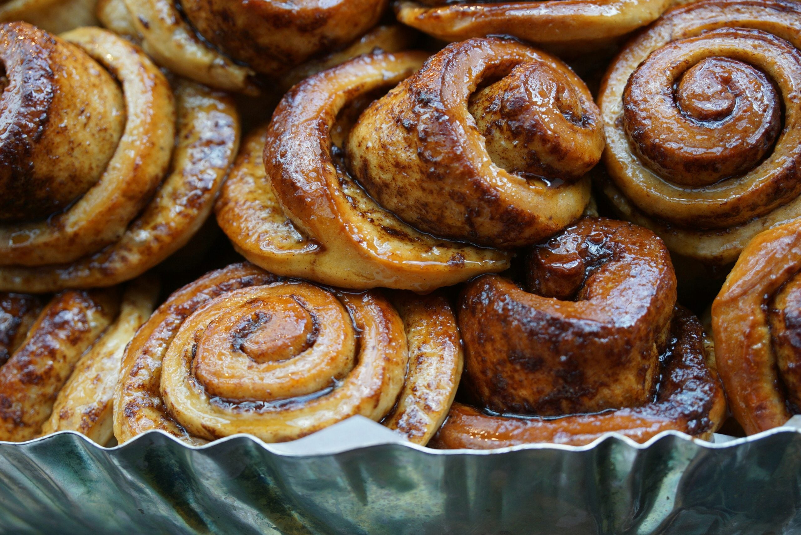 Freshly baked cinnamon rolls with a golden, glossy finish are closely packed together in a metal baking tray, showing swirls of cinnamon filling and a sticky, caramelized surface.