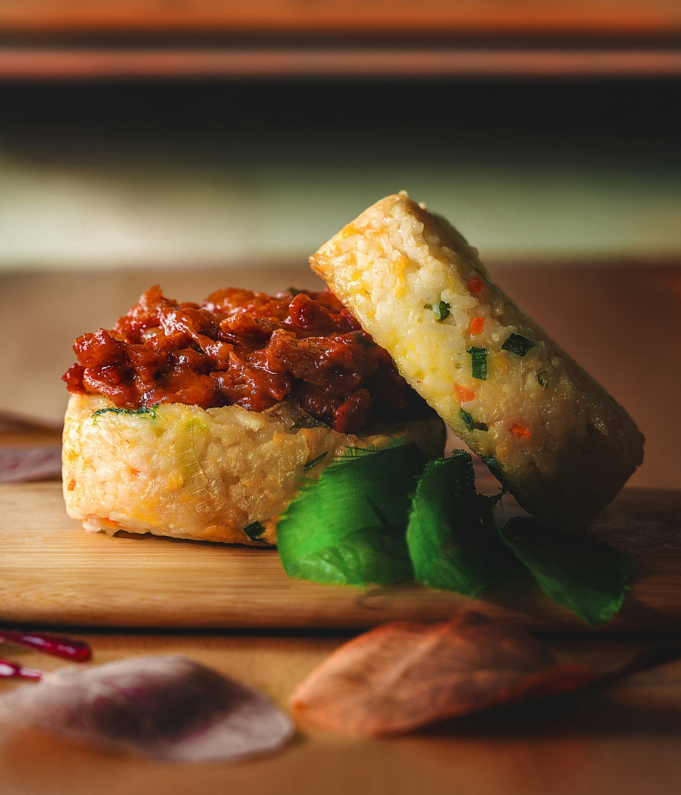 A gourmet burger with a filling of saucy, seasoned meat between two thick rice patties, served on a wooden board with fresh green leaves and decorative vegetable slices.
