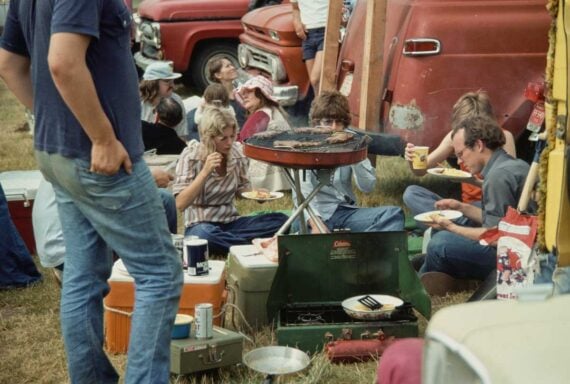 A group of people sit on the grass around a portable grill and stove, eating food at an outdoor gathering with coolers and vintage trucks in the background. Some are cooking while others eat and chat.