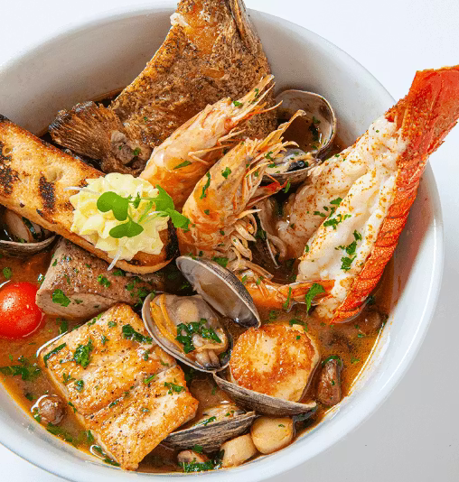 A bowl of seafood stew with fish, shrimp, lobster tail, clams, scallops, grilled bread, and vegetables, garnished with chopped herbs and served in a white bowl.