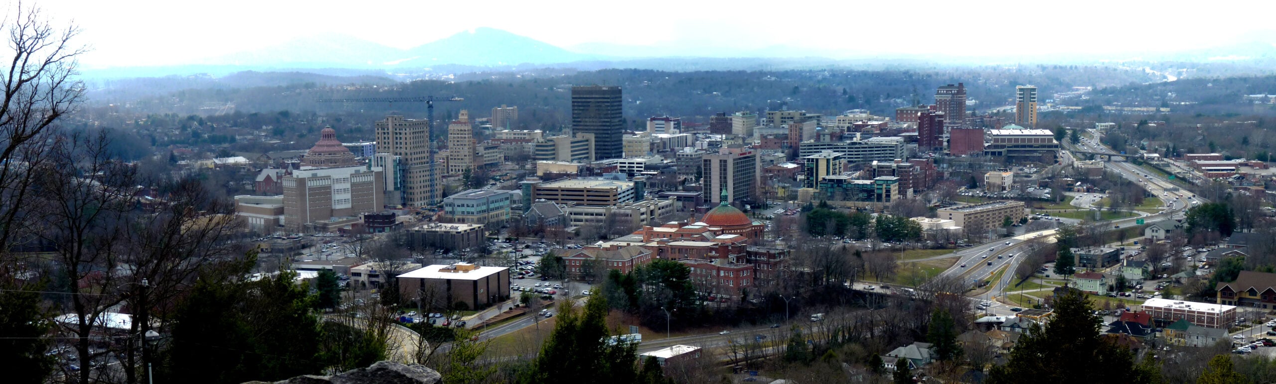 A panoramic view of a mid-sized city with a mix of modern and historic buildings, roads, and green spaces, surrounded by forested hills and distant mountains under a cloudy sky.
