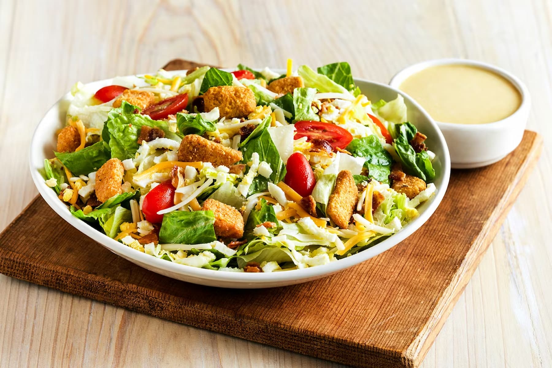 A bowl of fresh salad with lettuce, grape tomatoes, shredded cheese, and crispy croutons sits on a wooden board, with a small dish of creamy dressing beside it.