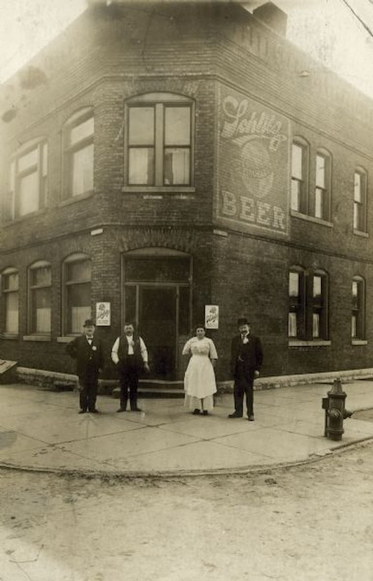 Four people stand outside a two-story brick building with "Schlitz Beer" painted on the wall. The group includes three men in suits and a woman in a white dress, posing on a street corner in front of the entrance.