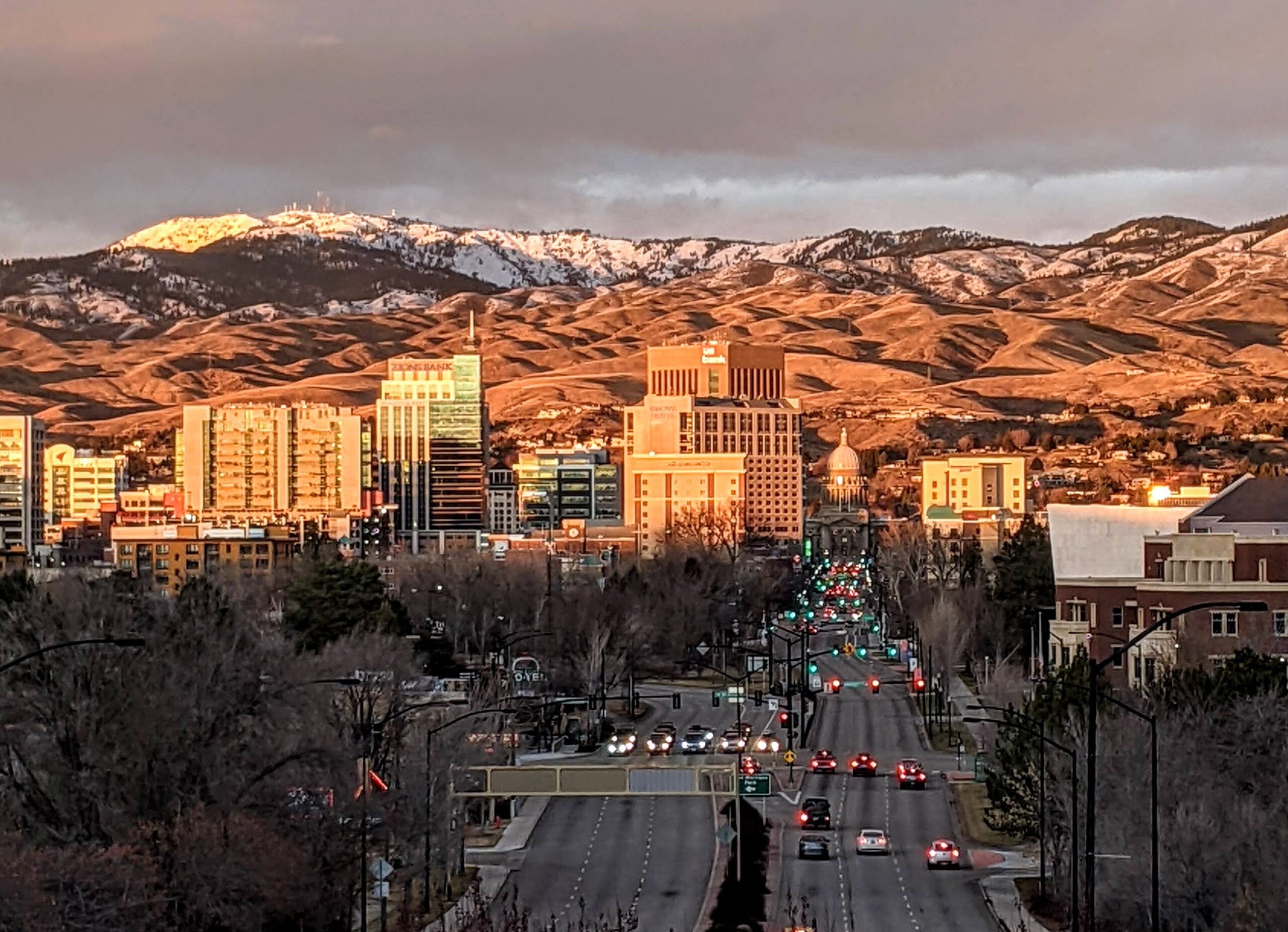 A cityscape at sunset shows tall buildings and a busy street with green traffic lights, set against rolling hills and snow-capped mountains under a cloudy sky.