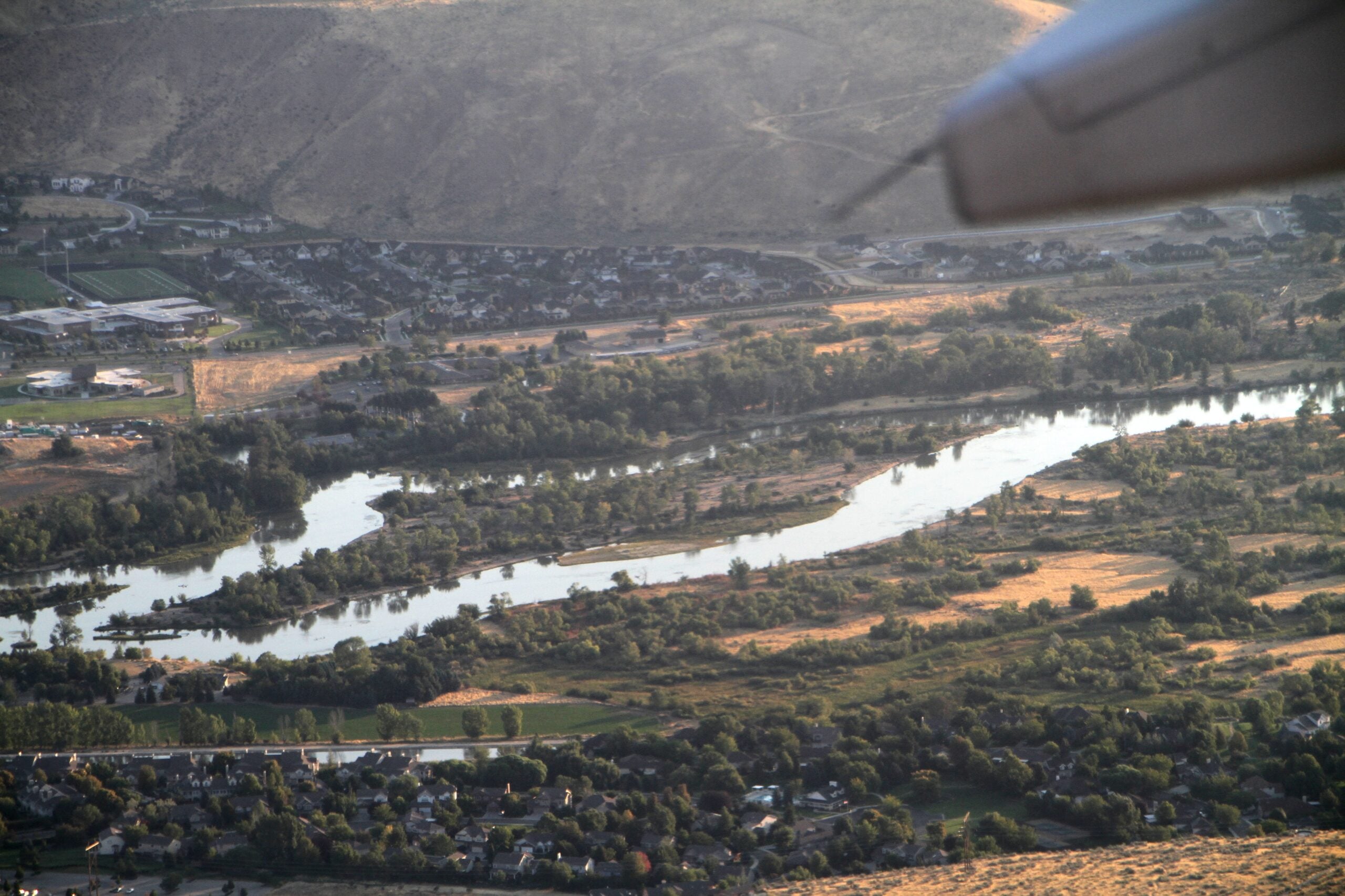 Aerial view of a winding river bordered by trees and small residential areas, with a dry hillside in the background and part of an airplane wing visible in the top right corner.