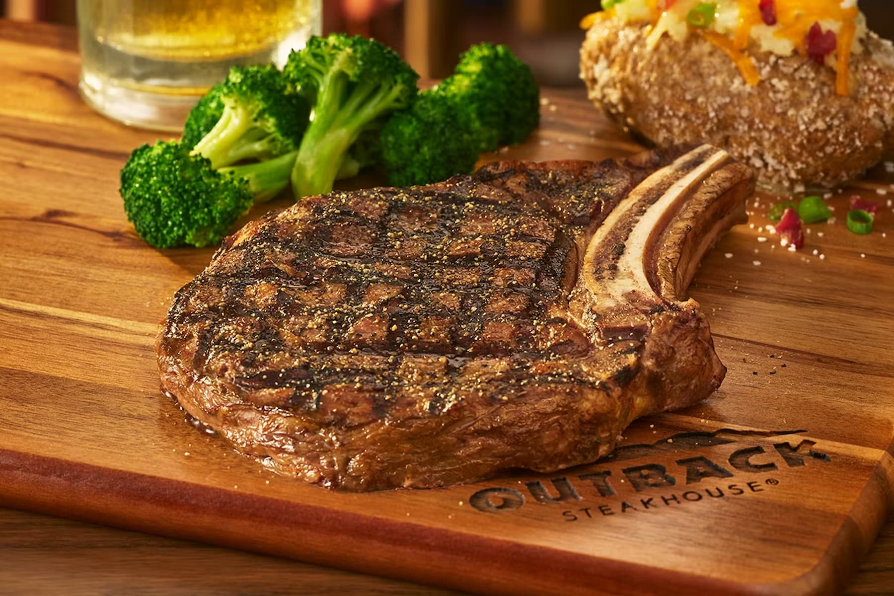A grilled bone-in steak with char marks sits on a wooden board labeled "Outback Steakhouse," served with broccoli and a baked potato topped with cheese, bacon, and scallions.