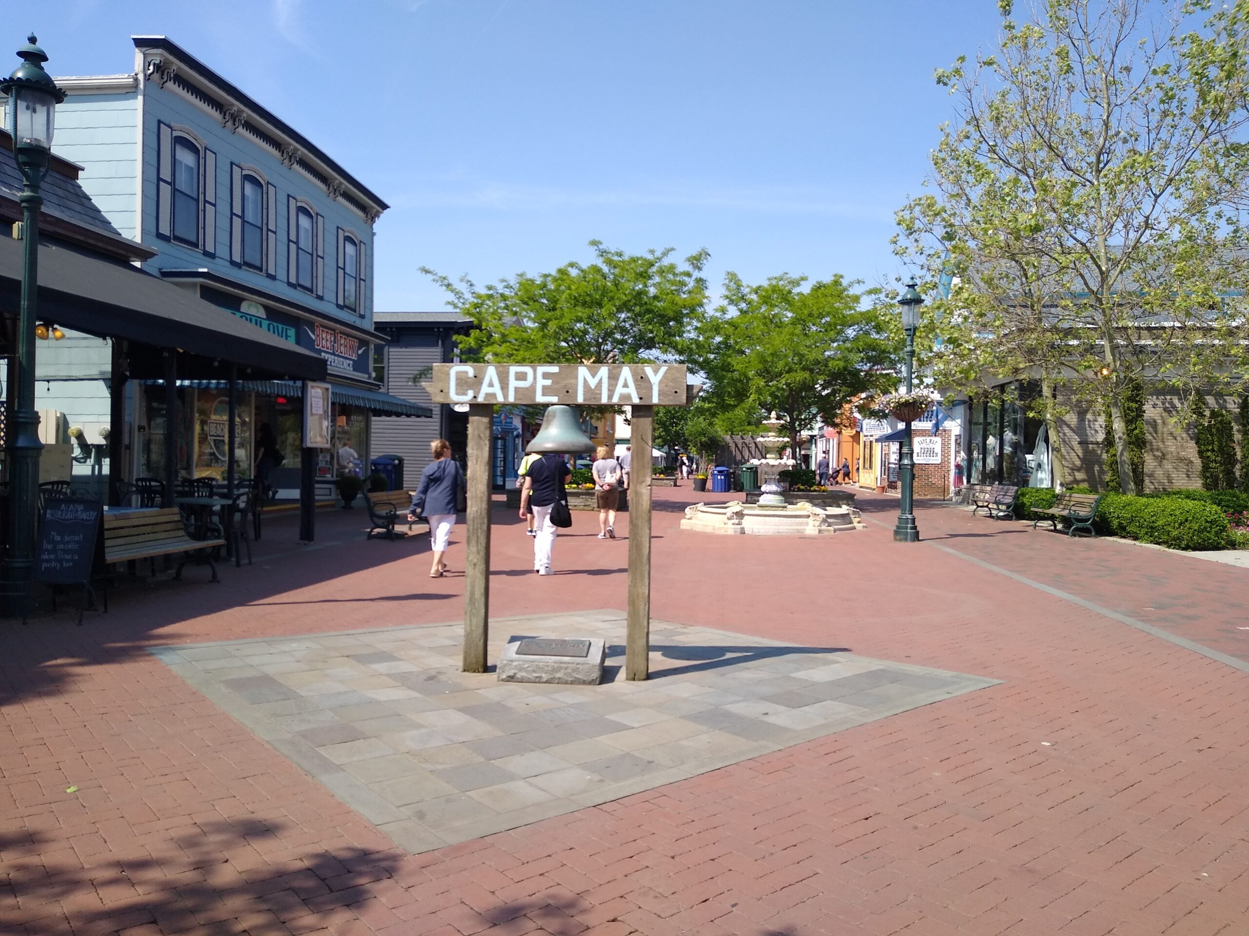 A sunny pedestrian street in Cape May features a large sign with a bell, shops on both sides, two trees, and people walking along the brick pathway.