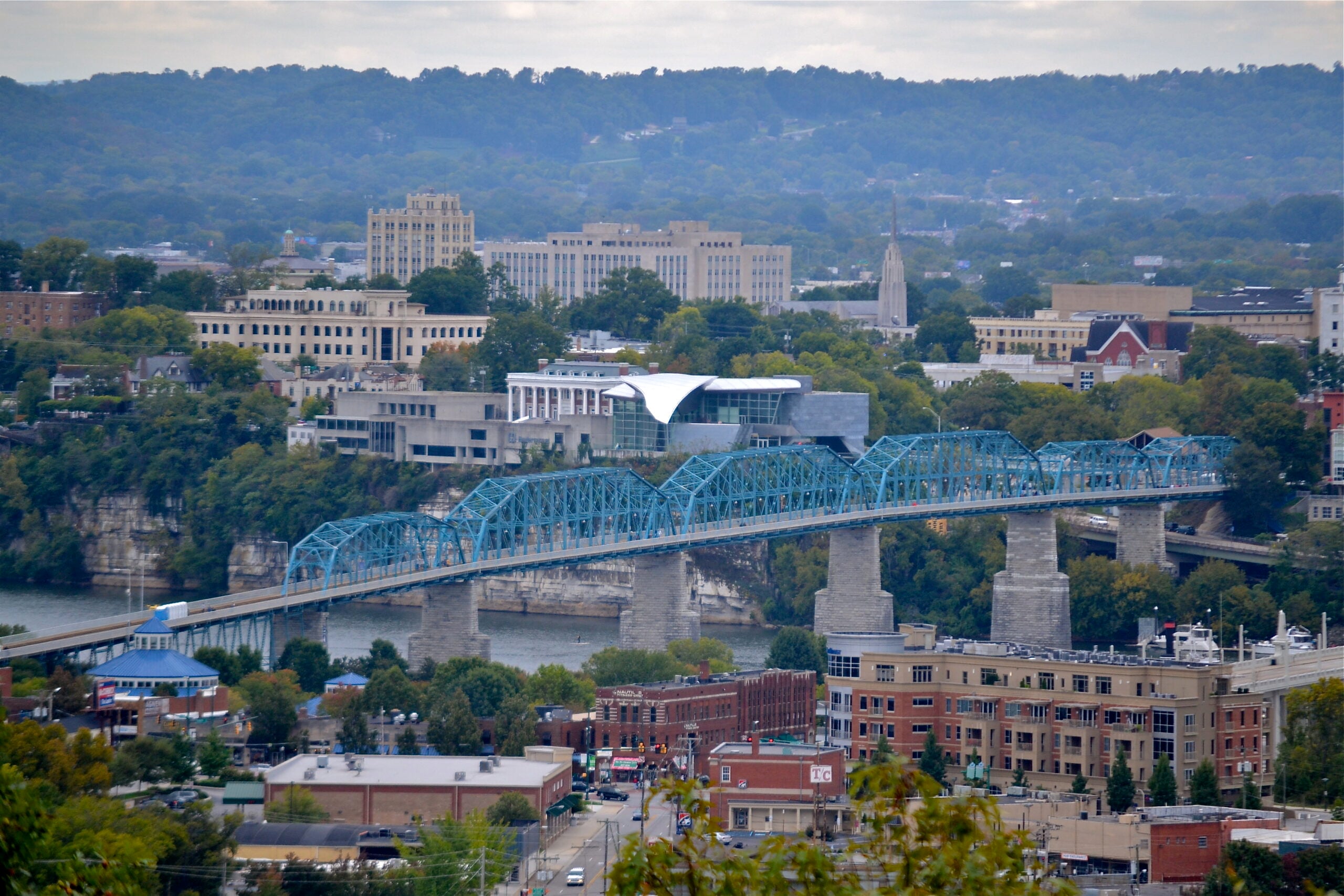 A blue truss pedestrian bridge crosses a river in a city with various buildings, trees, and hills in the background under a cloudy sky.