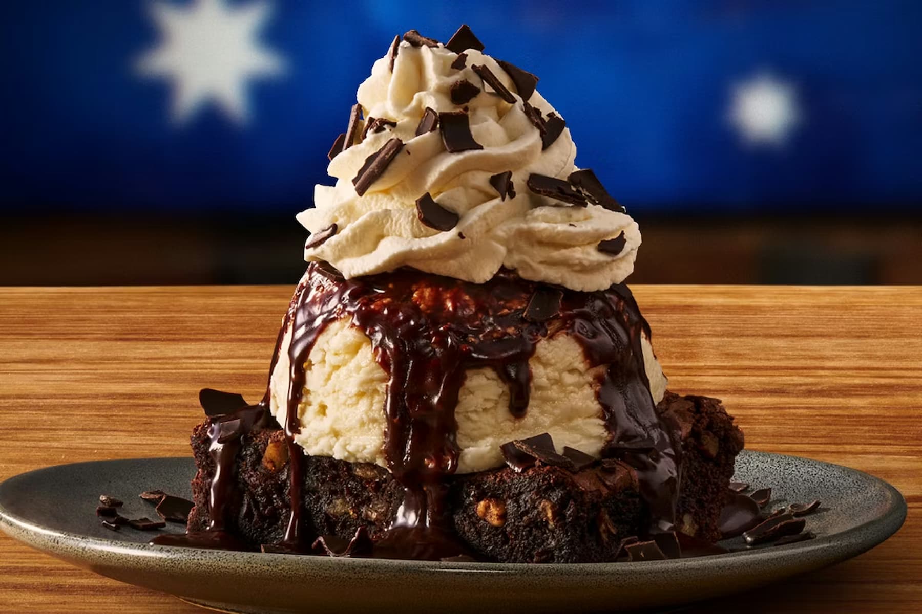 A dessert featuring a brownie base topped with a large scoop of ice cream, whipped cream, chocolate sauce, and chocolate shavings, served on a dark plate with a blurred blue and white background.