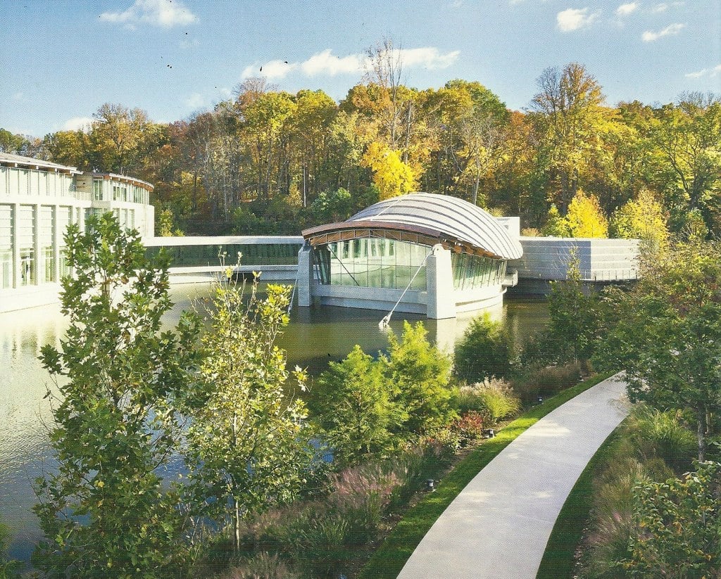 A modern building with curved roofs sits over a pond, surrounded by trees with autumn foliage. A curved walkway runs through landscaped greenery in the foreground under a sunny sky.