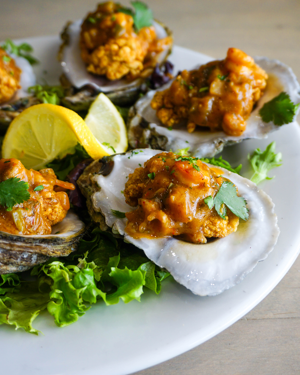 A white plate with oysters served on the half shell, topped with a savory orange sauce and garnished with cilantro, accompanied by leafy greens and lemon wedges.