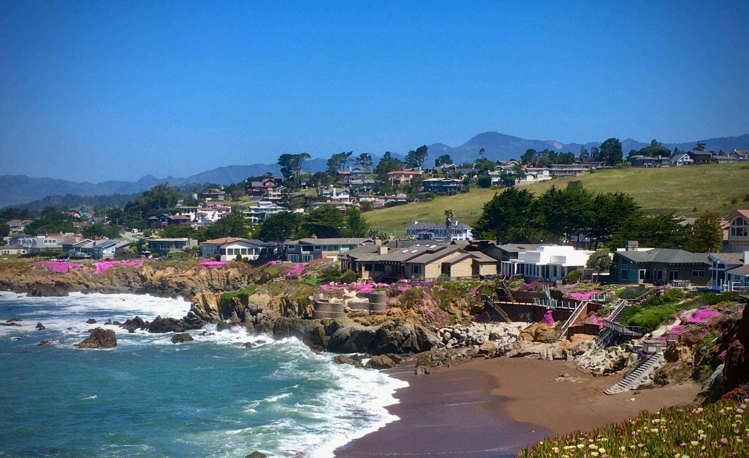 Coastal houses overlook a rocky shoreline with colorful pink flowers and waves crashing onto the sandy beach. Green hills rise behind the houses under a clear blue sky with distant mountains in the background.