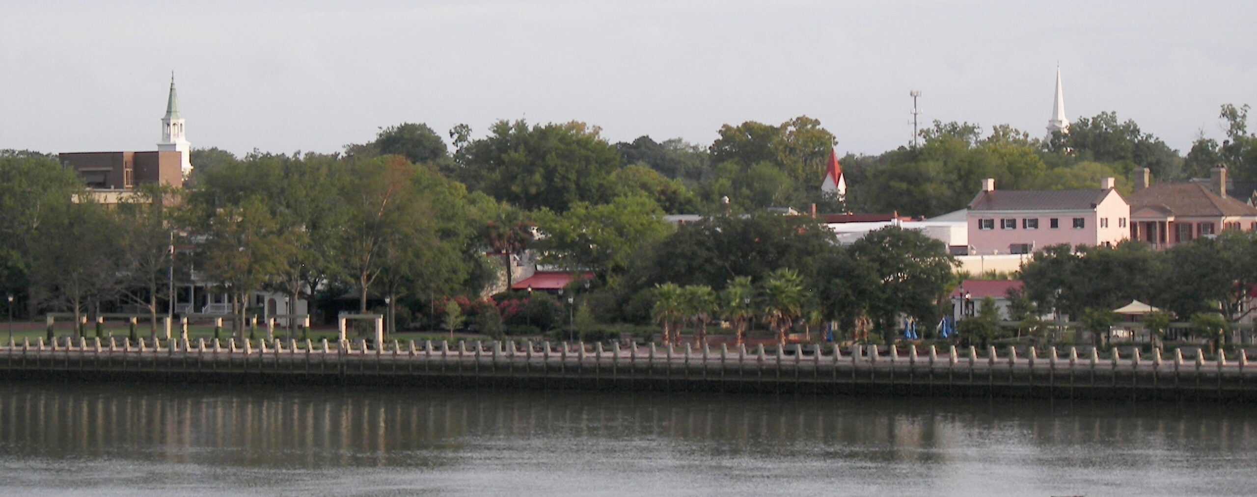 A calm river with a tree-lined waterfront, park benches, and buildings in the background; church steeples rise above the trees under a cloudy sky.