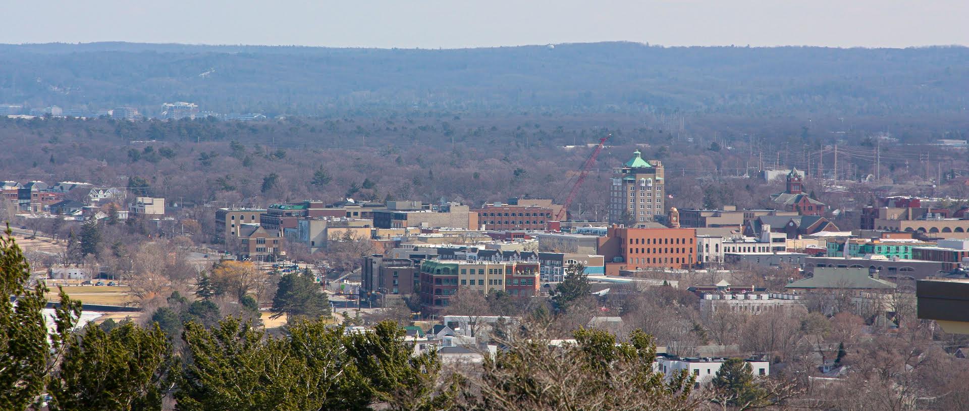 A wide view of a small city with low-rise buildings surrounded by trees, set against a backdrop of rolling hills under an overcast sky.