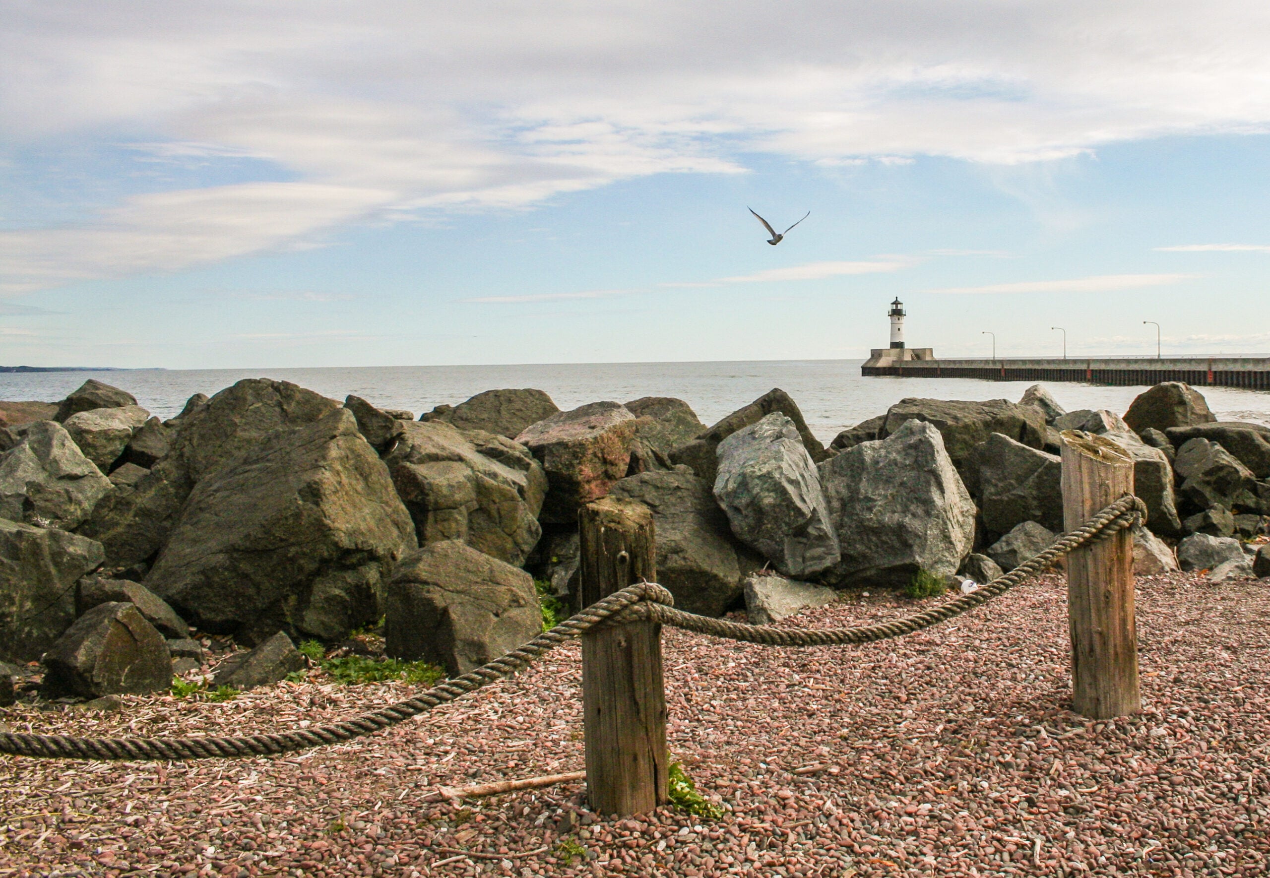 Rocky shoreline with large boulders in the foreground, a rope fence, and a distant pier with a lighthouse. A seagull is flying in the sky above the calm water under scattered clouds.
