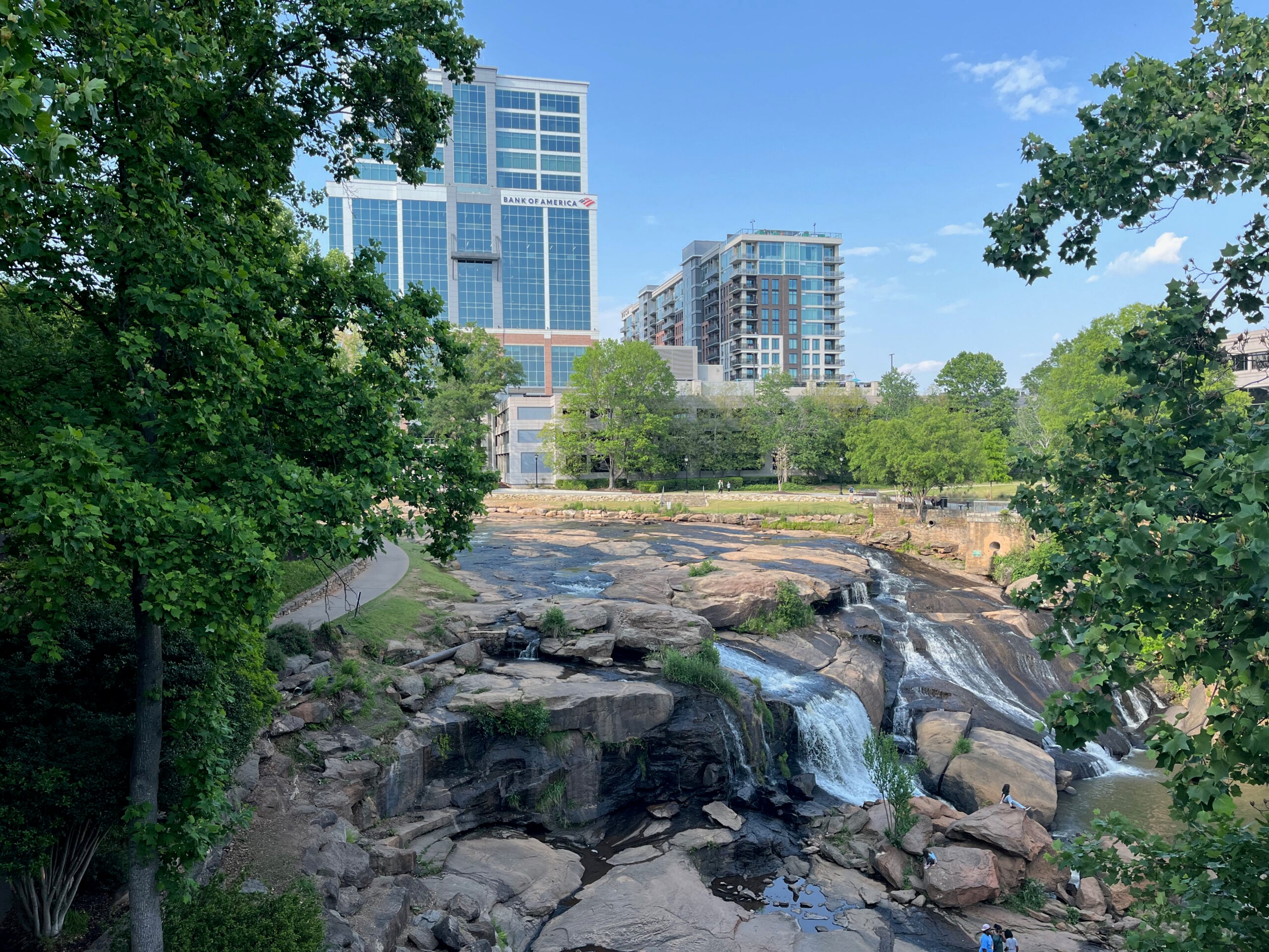 A scenic city park with a rocky waterfall in the foreground, surrounded by green trees, with modern high-rise buildings in the background under a blue sky.