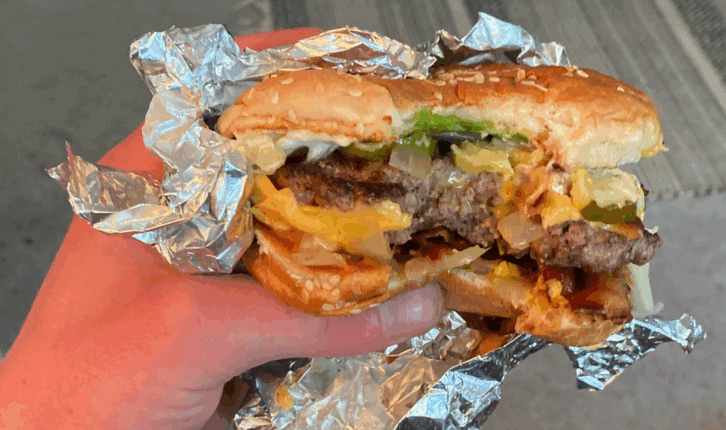 A hand holding a partially eaten cheeseburger wrapped in aluminum foil, showing lettuce, cheese, onions, and a beef patty inside a sesame seed bun.