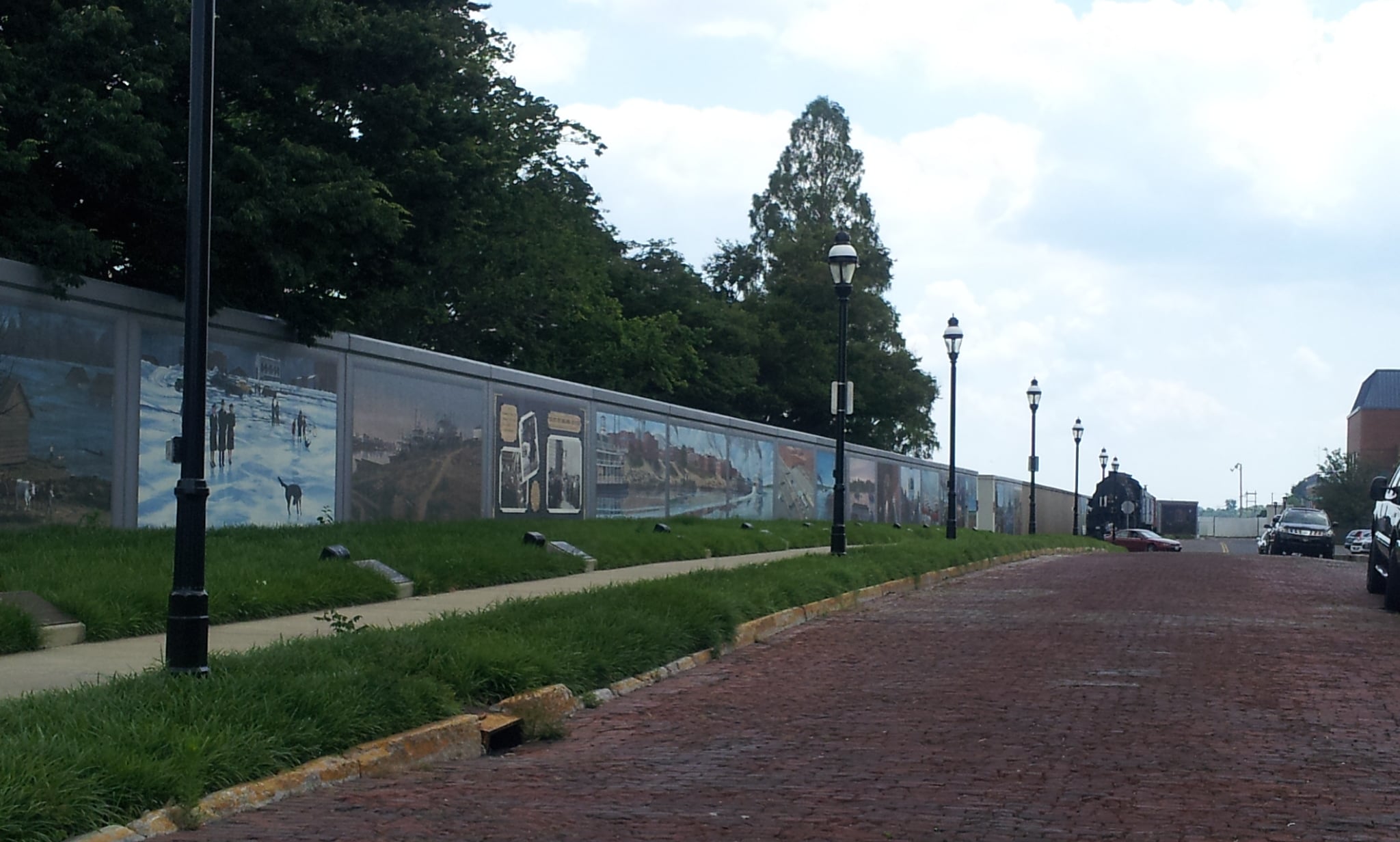 A brick-paved street runs alongside a mural-covered wall lined with black street lamps and grass. Trees and parked cars are visible on both sides under a partly cloudy sky.