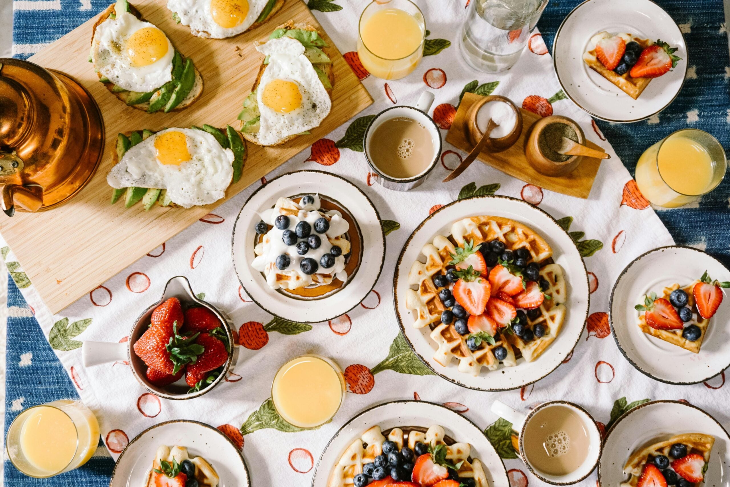 A breakfast spread on a table with waffles topped with strawberries and blueberries, avocado toast with fried eggs, glasses of orange juice, a pitcher of strawberries, coffee cups, and a teapot on a patterned tablecloth.