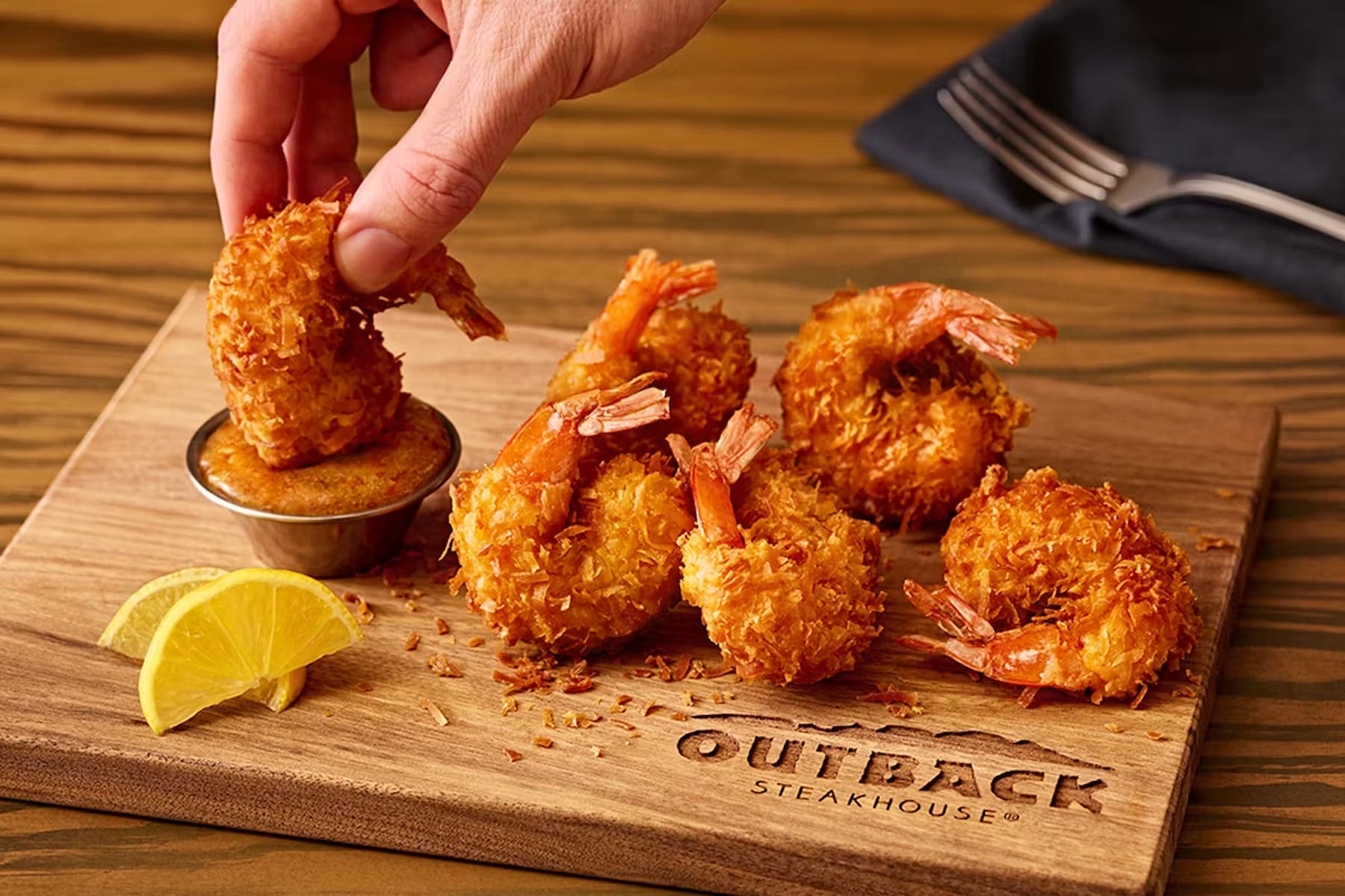 A hand dips a piece of crispy fried shrimp into a sauce cup on a wooden board with several coconut shrimp, lemon wedges, and the Outback Steakhouse logo. A fork and napkin are in the background.
