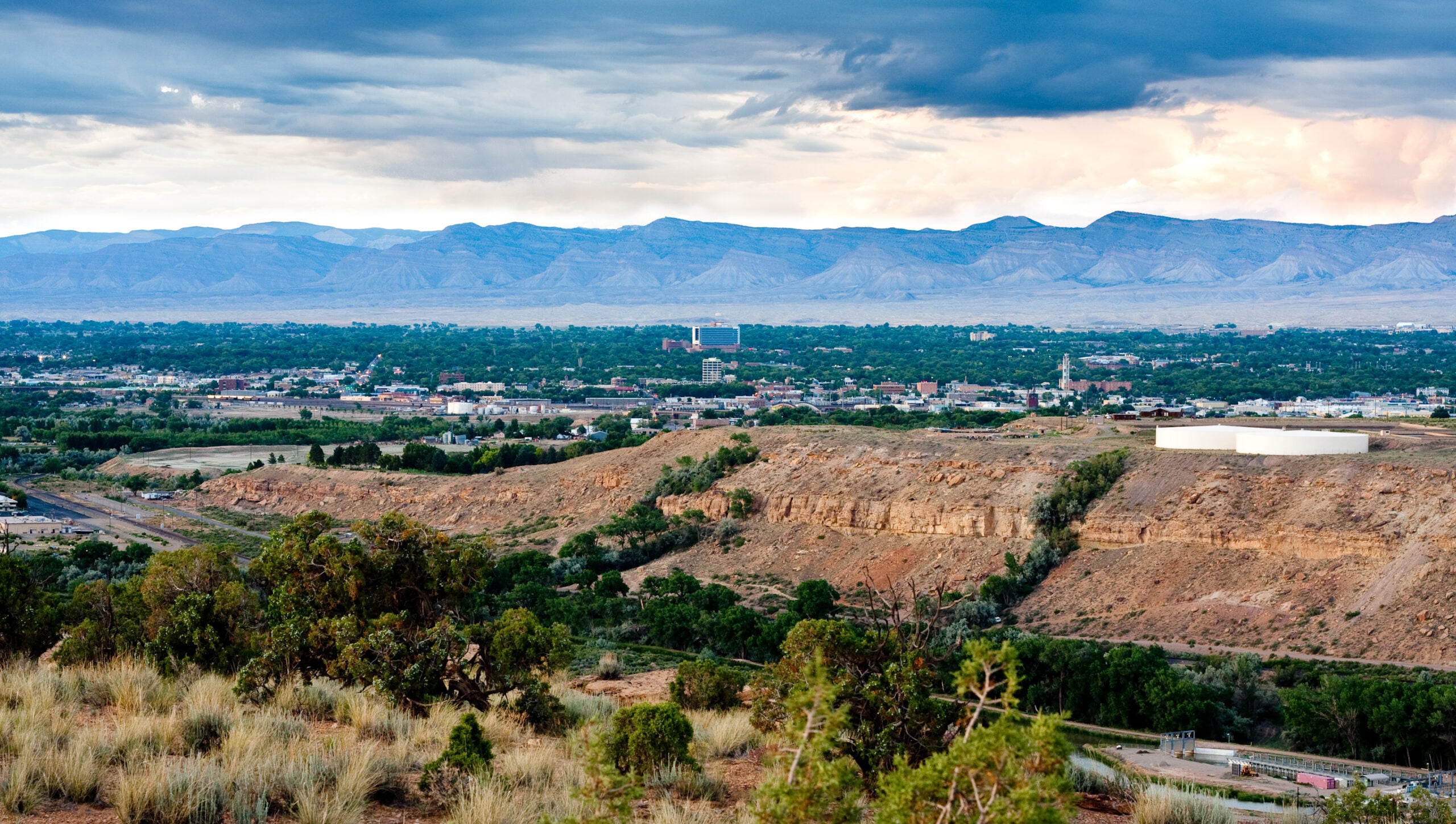 A panoramic view of a city surrounded by trees and hills, with a large, rocky plateau in the foreground and blue mountains in the distance under a partly cloudy sky.