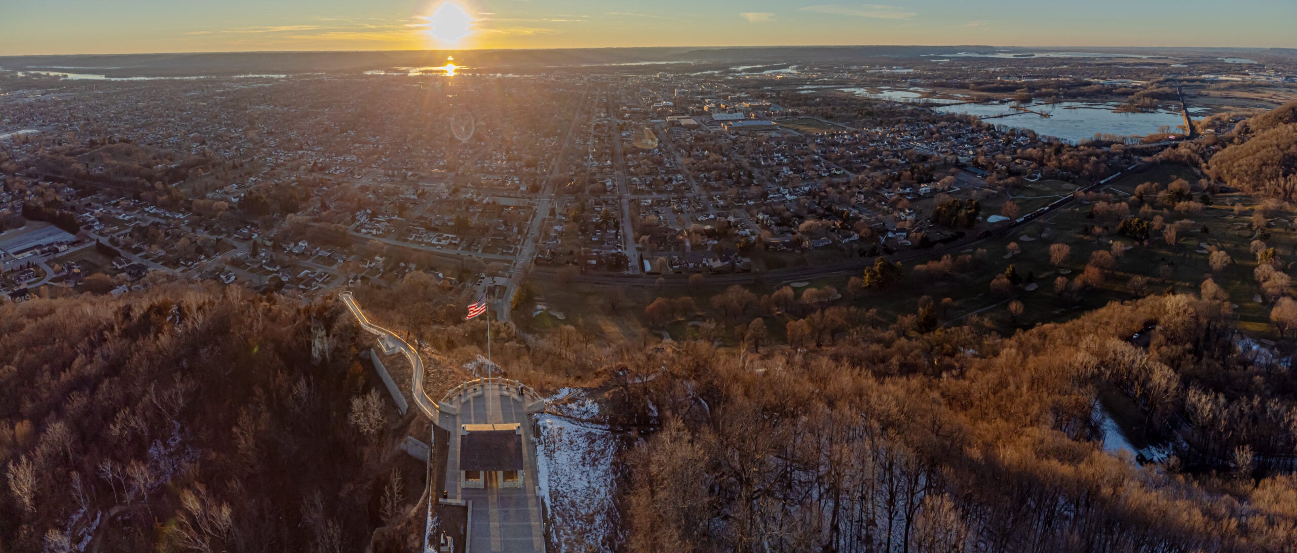 Aerial view of a city at sunset with a wide river, residential neighborhoods, and a prominent staircase or monument in the foreground on a wooded hill. The sun is low on the horizon, casting a warm golden light.