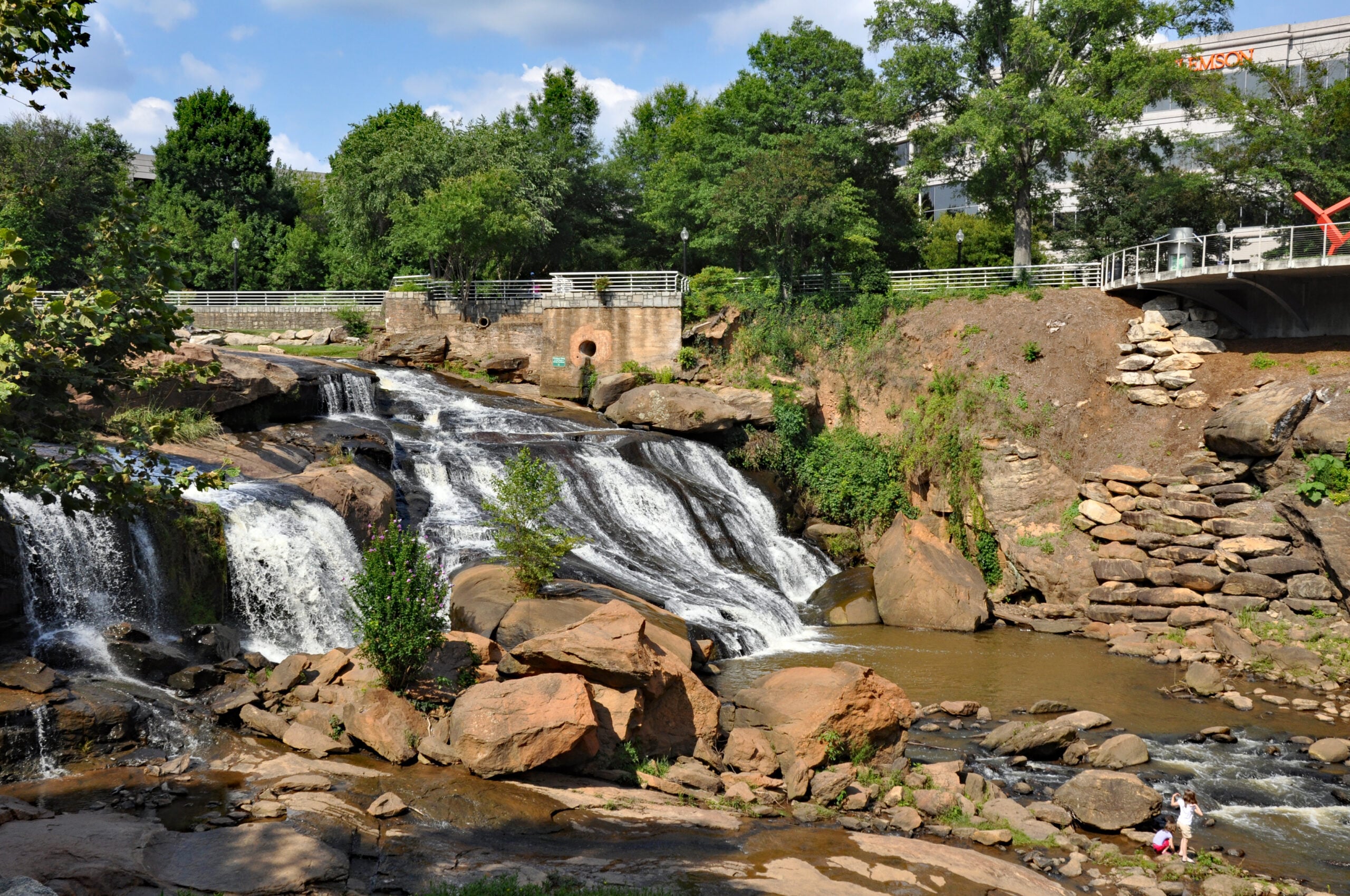 A mid-sized waterfall flows over rocks surrounded by trees and greenery. A pedestrian bridge and pathway are visible above the falls, with people walking nearby. A few buildings and sculptures appear in the background.