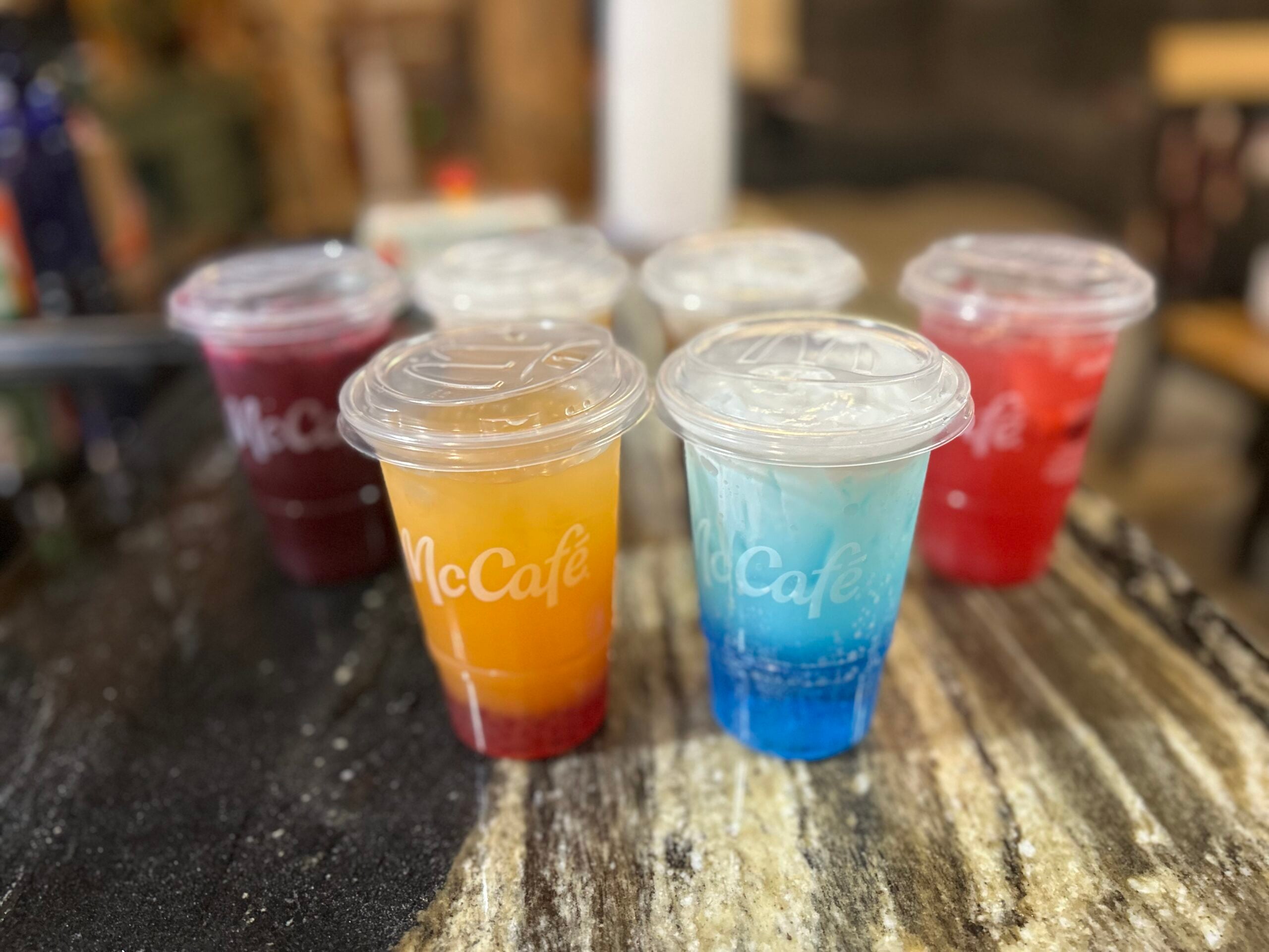 Six colorful McCafé drinks in clear plastic cups with lids, displayed on a dark marble surface. The drinks are various shades of orange, blue, red, and purple.