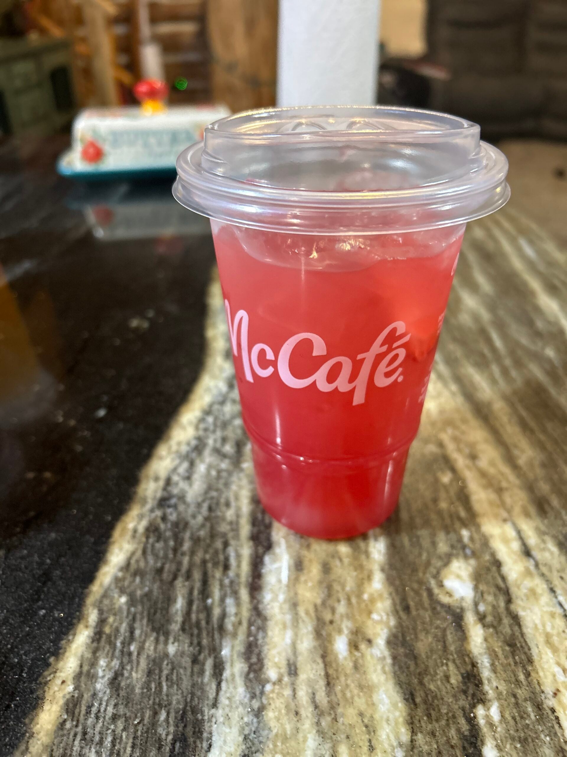 A McCafé plastic cup filled with a bright red, iced beverage sits on a dark, marbled countertop. The cup has a clear lid, and the background is slightly out of focus.