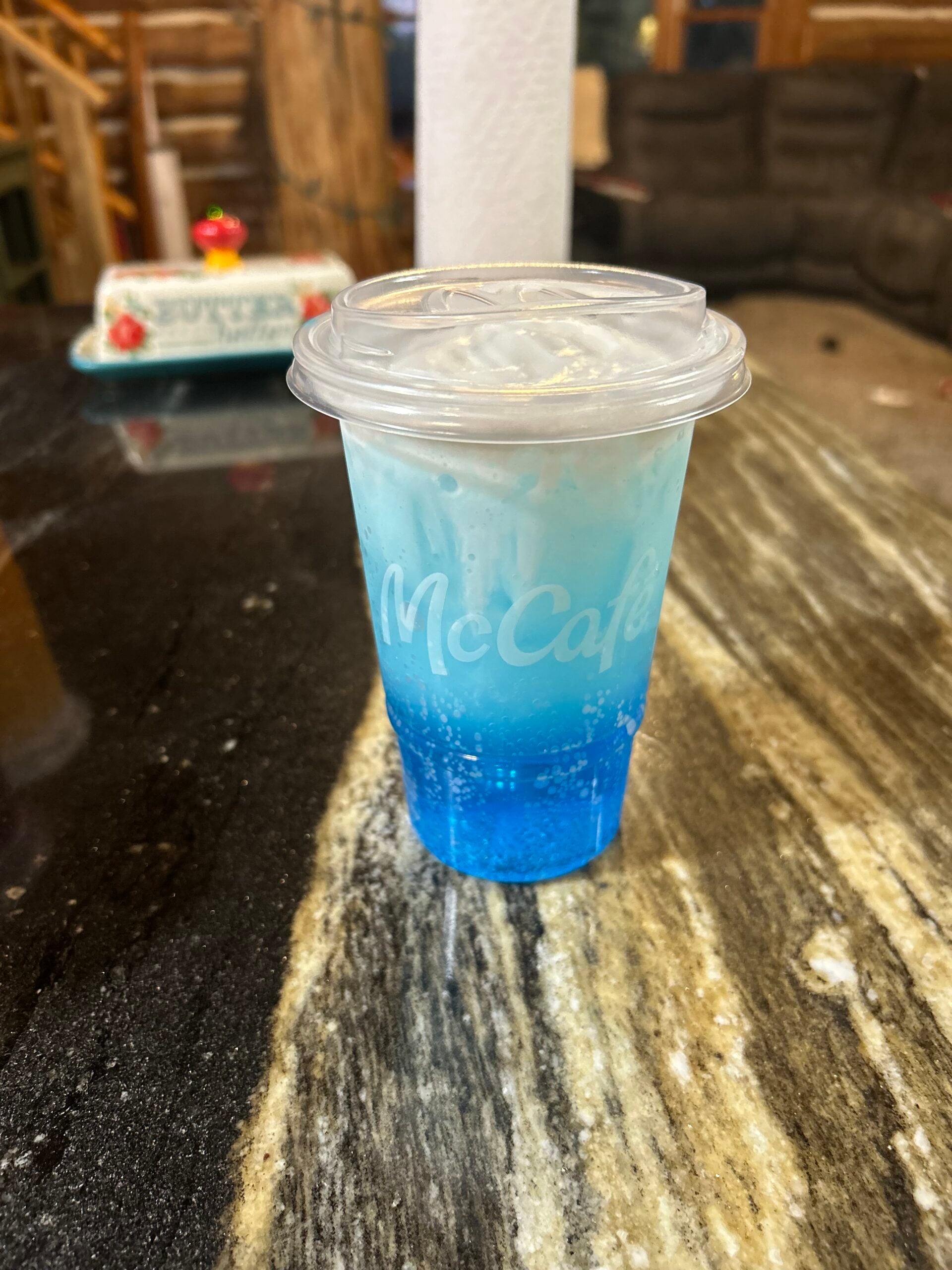 A clear McCafé cup filled with a blue beverage and ice sits on a marbled countertop. The drink appears fizzy, and the background shows a rustic indoor setting with a couch and blurred decor.