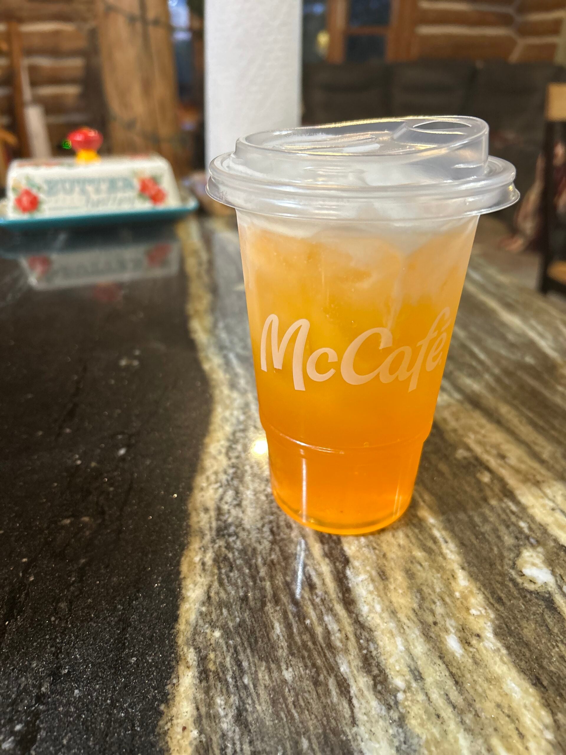 A clear McCafé cup with an orange iced drink and a plastic lid sits on a dark marble countertop, with a rustic wooden background and a decorated cake in the distance.