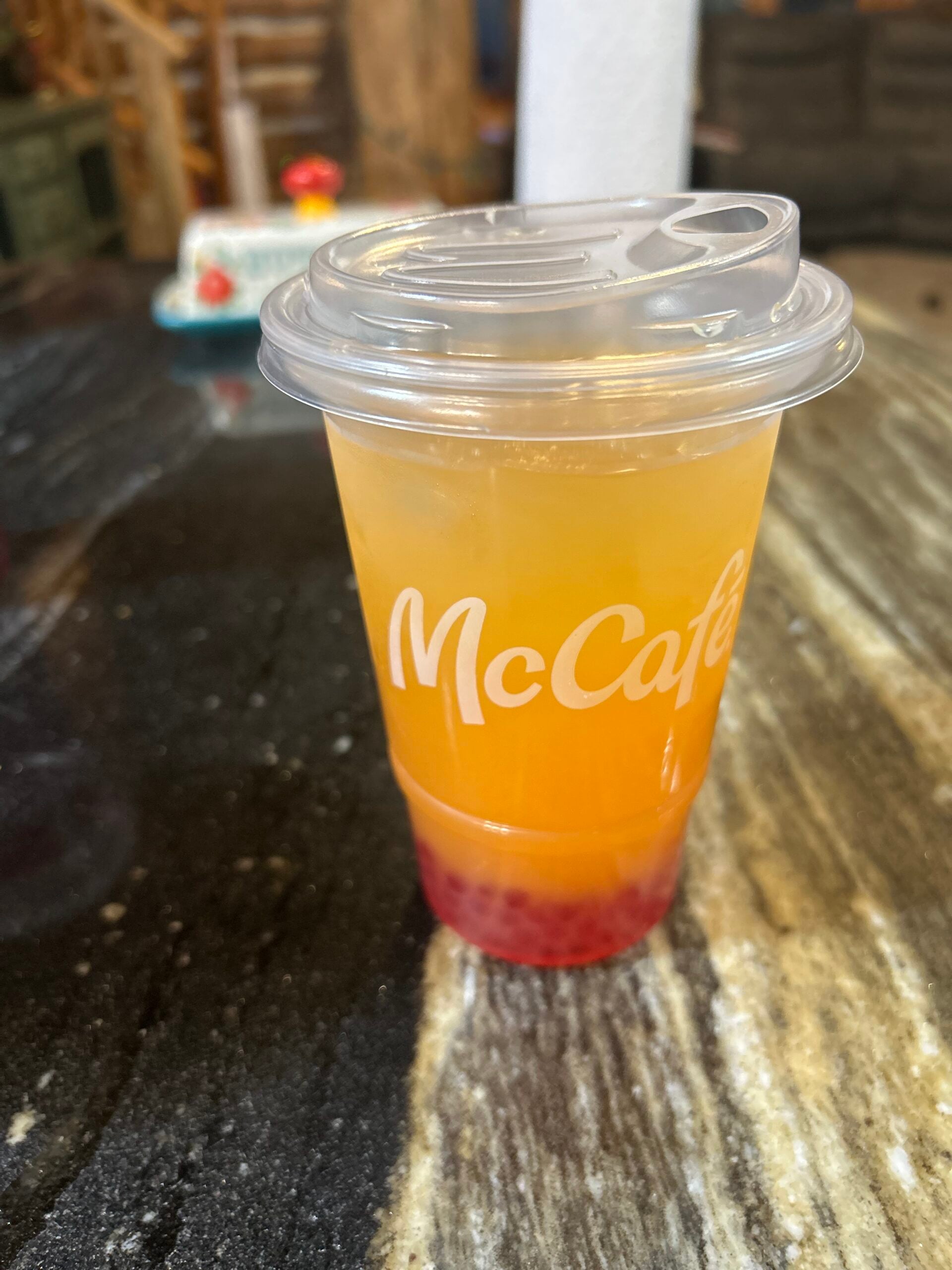 A plastic McCafé cup with an orange and yellow layered drink, topped with a clear lid, sits on a marbled table. Red boba pearls are visible at the bottom of the cup.